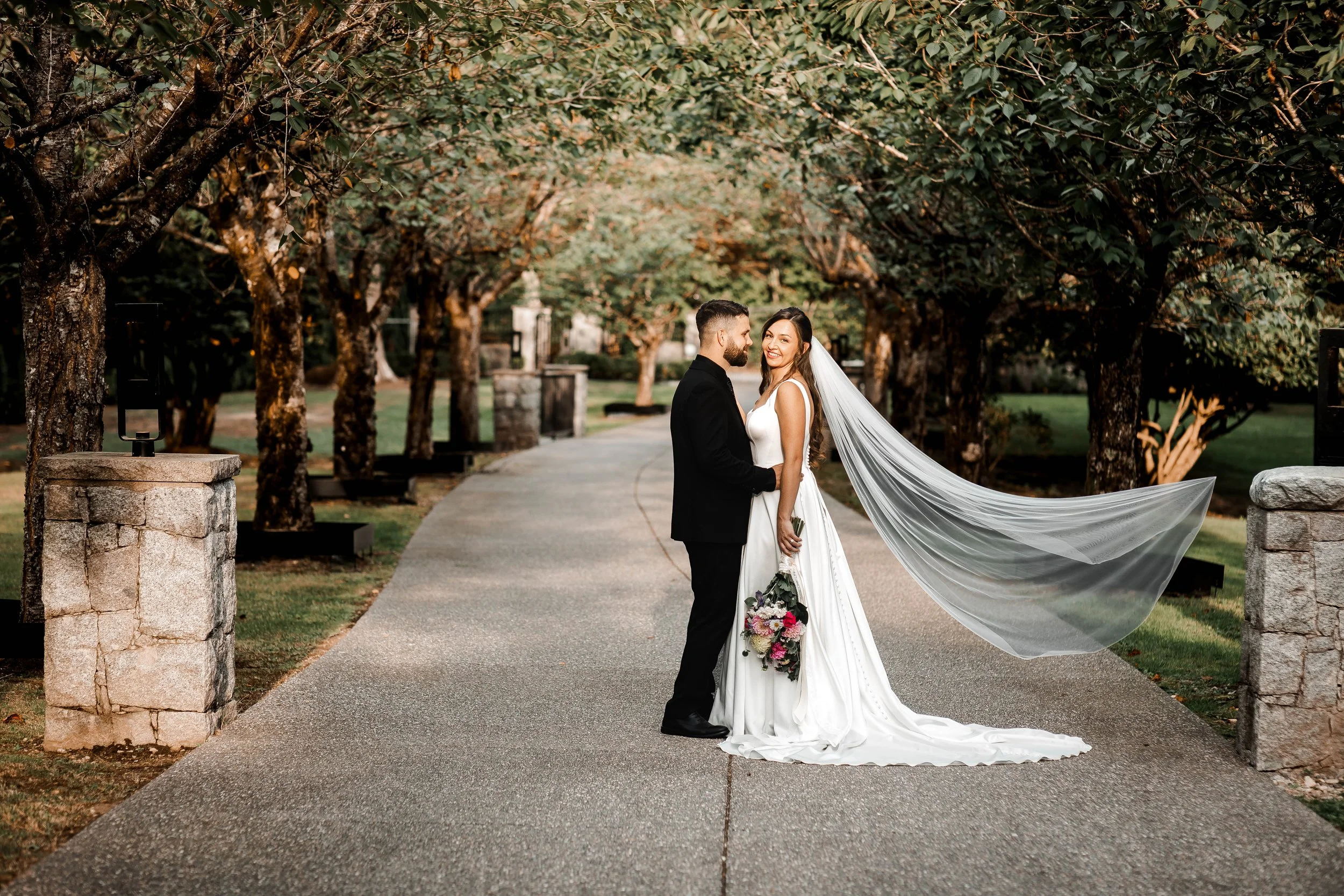 Bride and groom posing on a tree-lined pathway as the bride's long veil flows behind her during romantic wedding portraits.