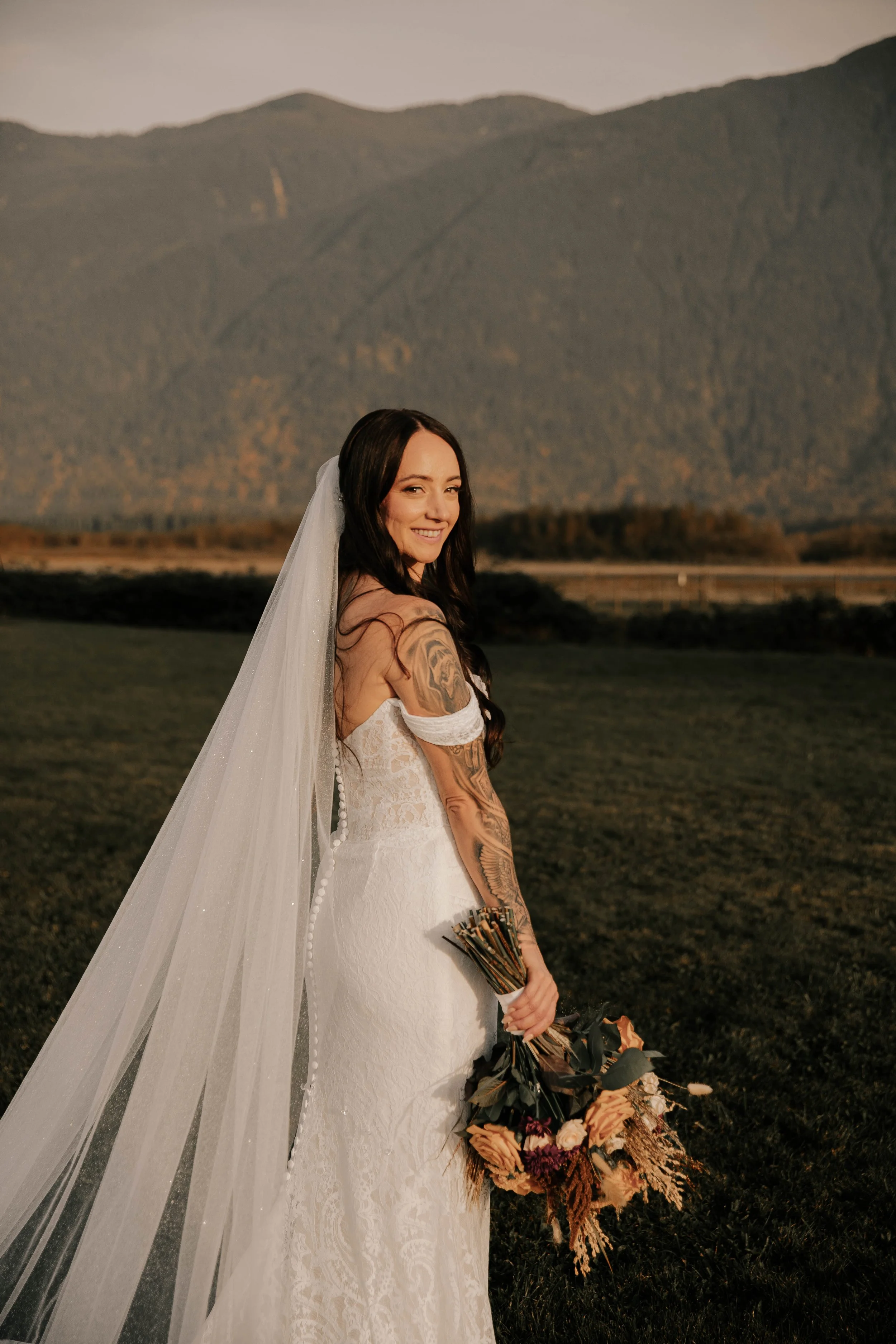 Bride in a flowing white wedding gown and veil holding a rustic bouquet against the mountain backdrop at Fraser River Lodge in Agassiz, BC.