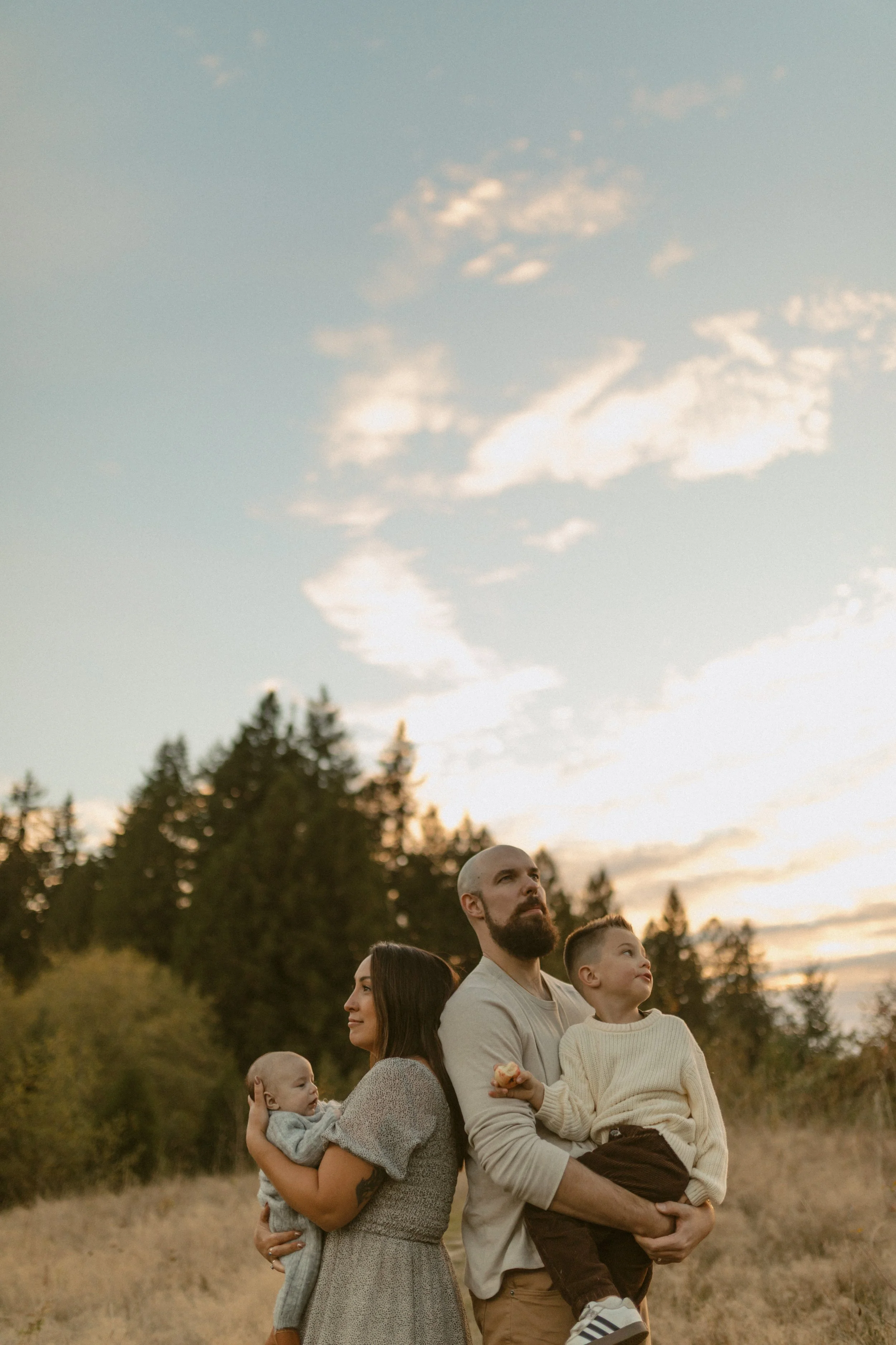 A family of four, including two adults and two children, stands outdoors in a field with tall grass and trees in the background, during sunset, looking upwards.
