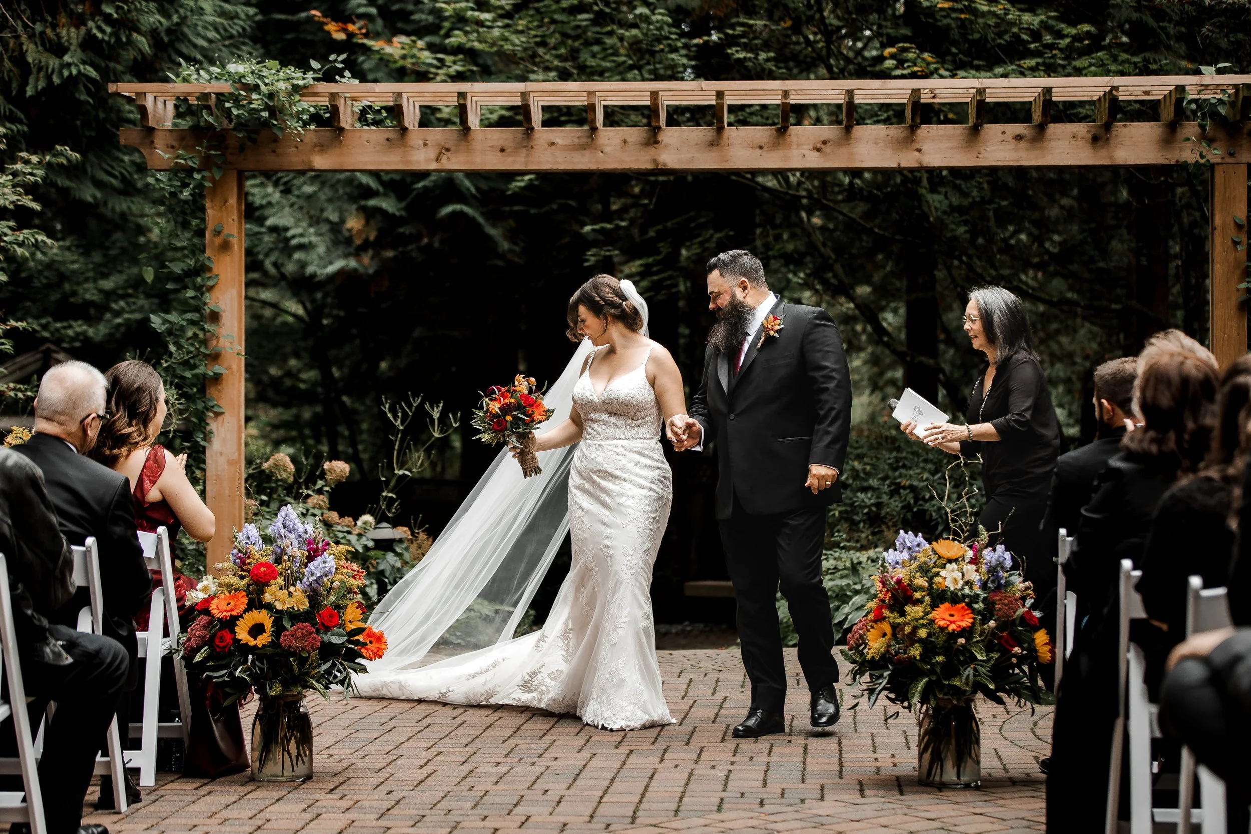 Bride and groom walking hand in hand beneath a rustic wooden pergola during their outdoor wedding ceremony at Whonnock Lake in Maple Ridge, BC.