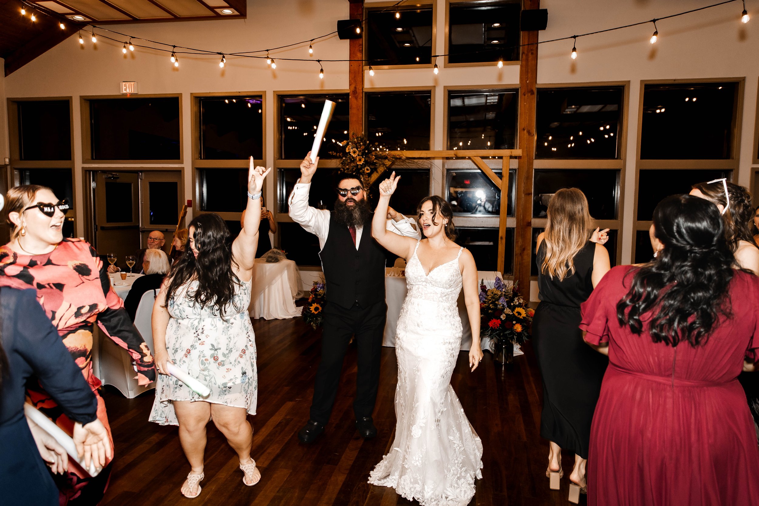 A wedding reception with guests dancing and celebrating, including a bride and groom in the center. The bride is in a white lace wedding dress, and the groom is in a black vest and trousers. The setting is indoors with string lights hanging from the ceiling and large windows in the background, creating a festive atmosphere.