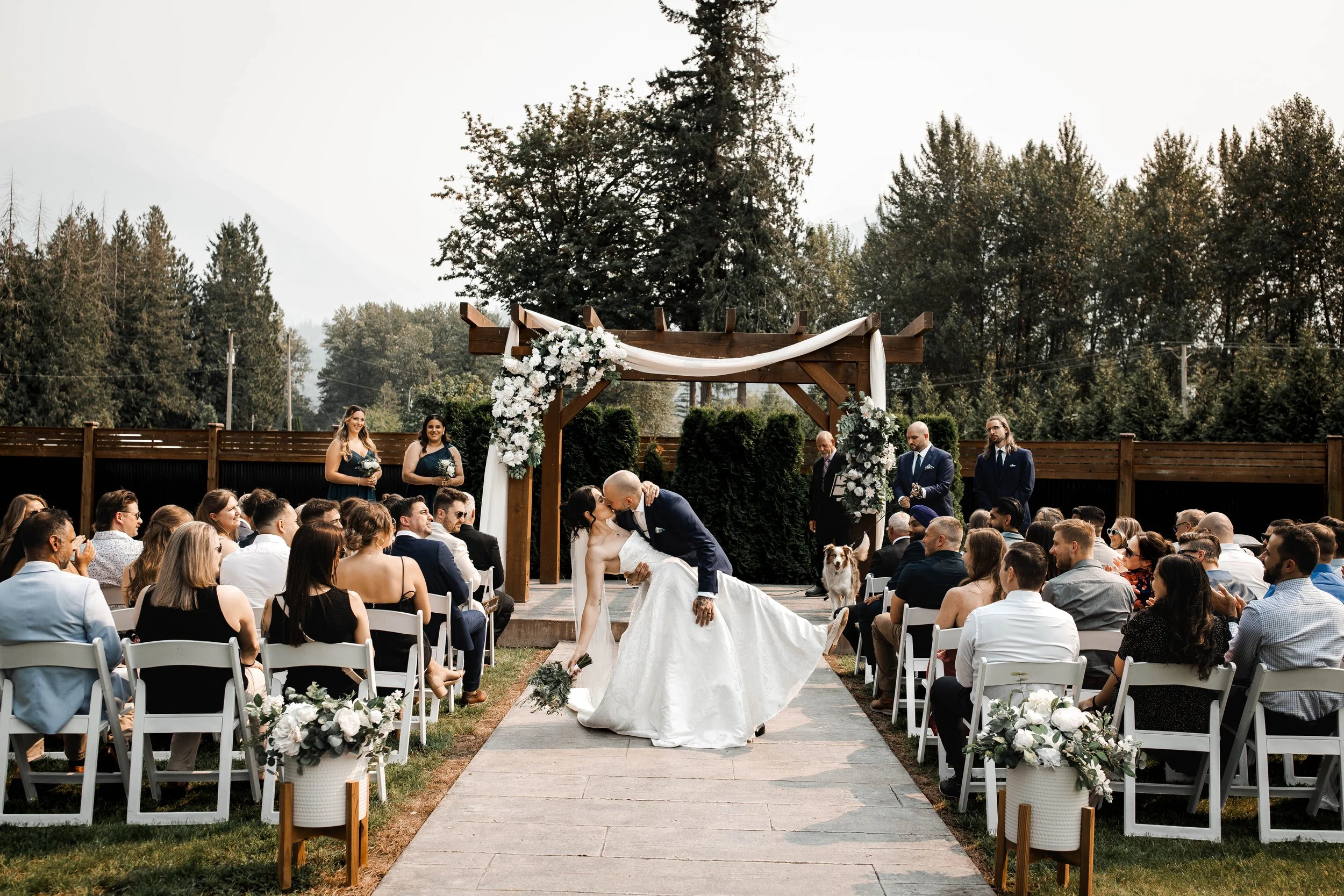 Bride and groom sharing a romantic first kiss during their outdoor wedding ceremony at Bridlewoods in Chilliwack, BC, surrounded by guests and mountain scenery.