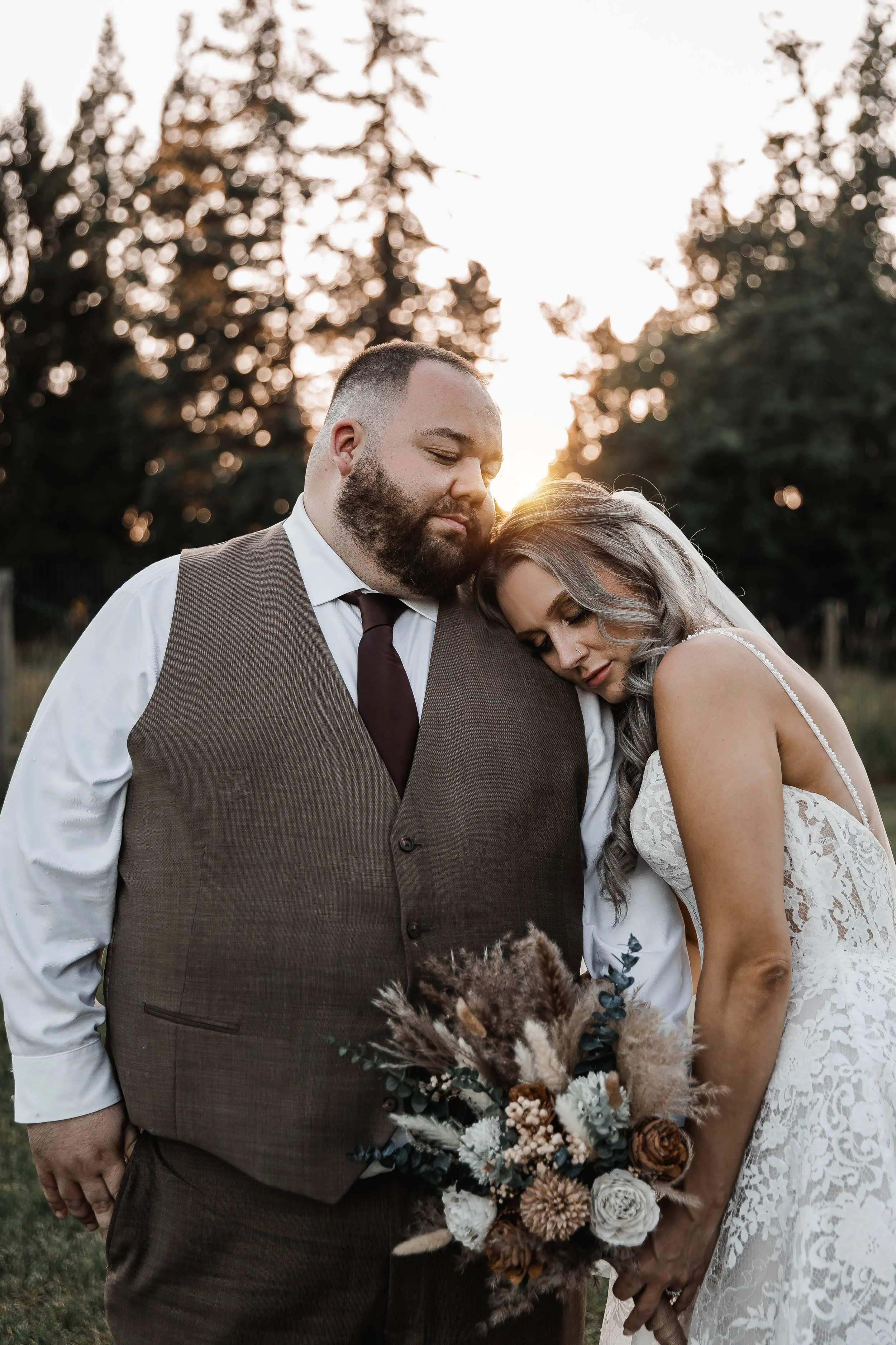 Bride resting her head on the groom’s shoulder while holding a rustic wedding bouquet outdoors at sunset