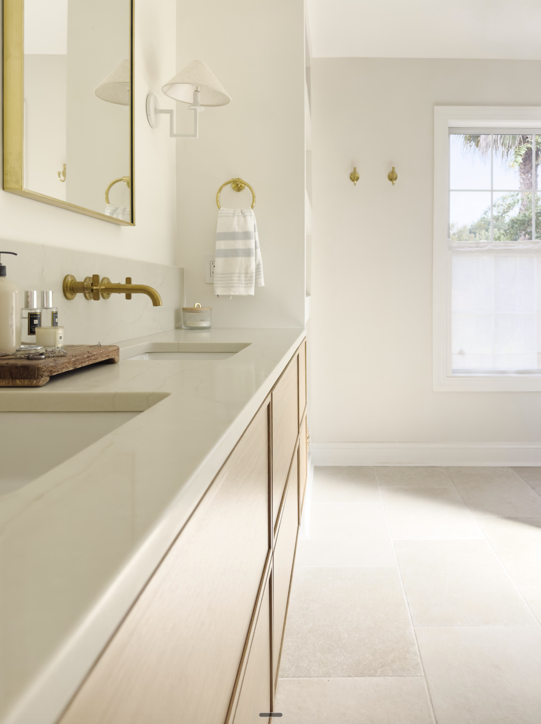 A bathroom with a beige countertop, wooden cabinets, gold fixtures including a faucet, towel ring, and wall hooks. There are toiletries on the counter, a mirror above the sink, a wall-mounted lamp, and a window showing an outdoor view with trees.