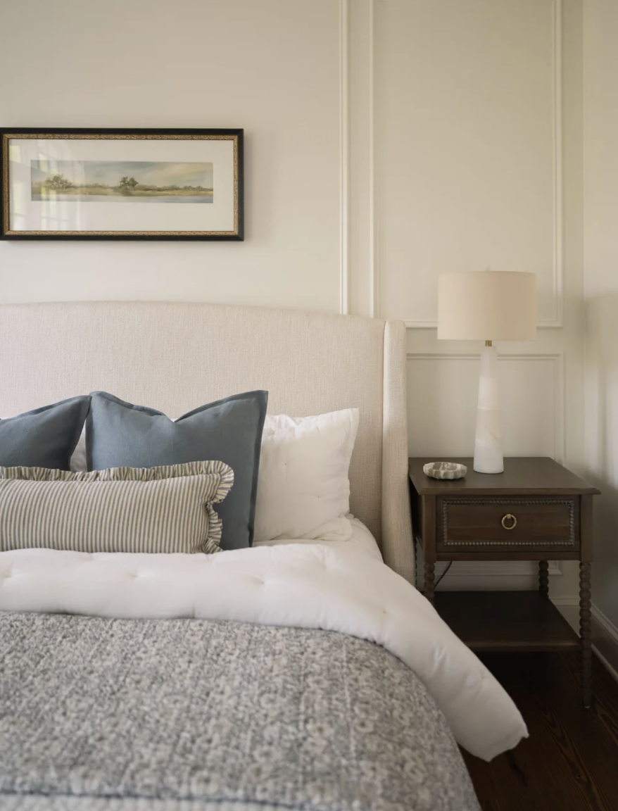 A bedroom corner with a beige upholstered bed, blue and striped pillows, a wooden nightstand with a white table lamp, a decorative stone, and a framed landscape painting above the headboard.