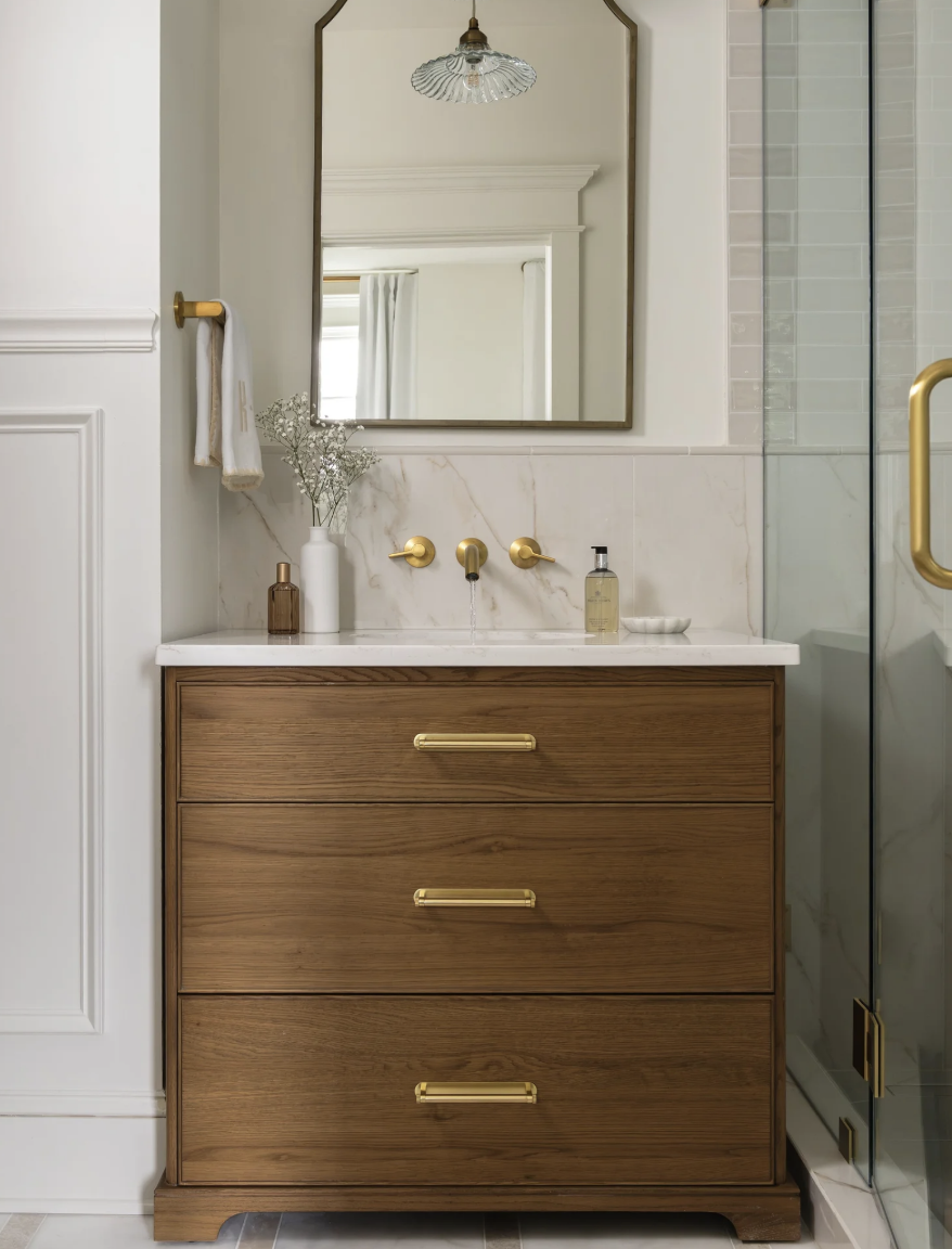 Modern bathroom vanity with a white marble countertop, gold handles, and a large mirror. Decor includes a vase with white flowers, soap dispenser, and a small dish. The background shows a glass shower enclosure with gold hardware.