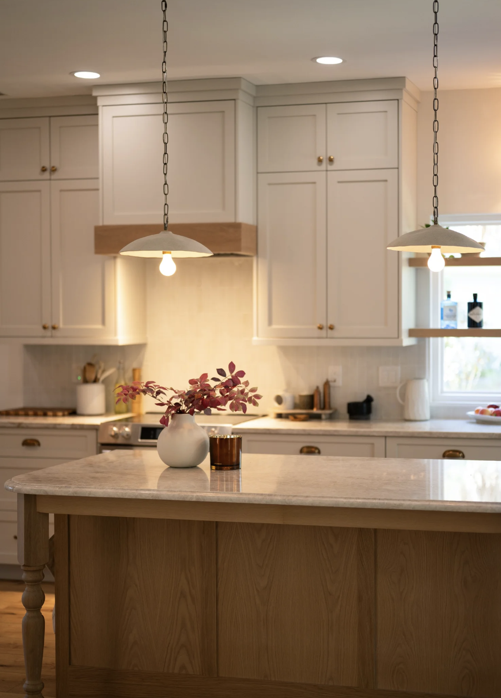 A cozy kitchen with cream cabinets, pendant lighting, and a marble island table. A white vase with red and purple leaves and a small amber candle sit on the kitchen island. The background includes a stove, countertop, kitchen utensils, and a window w