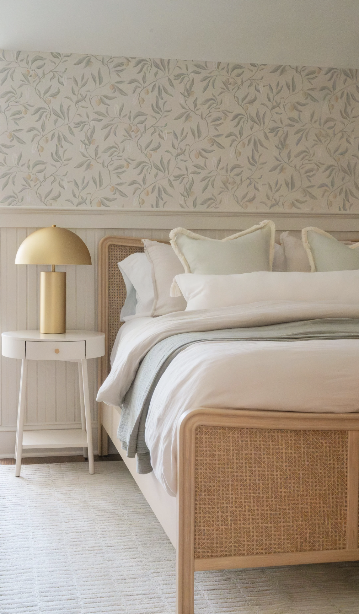 A neatly made bed with white and light green pillows, a white duvet, and a beige bed frame with woven detail, next to a white nightstand with a gold lamp, against a wall with floral wallpaper.