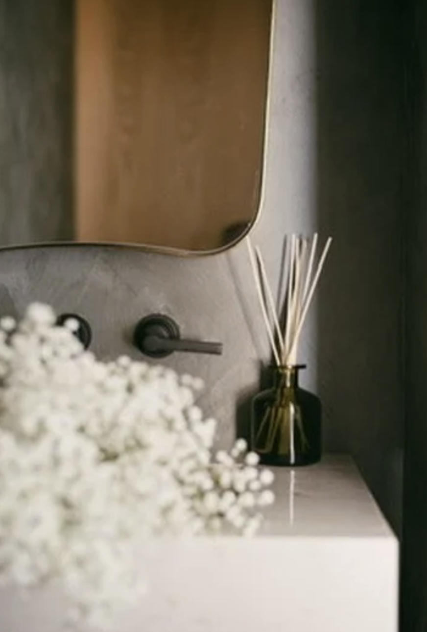 A decorative home interior with a mirror, a black reed diffuser, and white flowers on a marble surface.