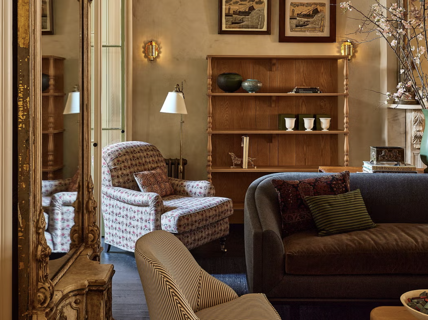 A cozy living room featuring a floral armchair, a dark gray sofa with decorative pillows, a wooden bookshelf with vases and books, a large mirror with an ornate gold frame, and framed artwork on the walls.