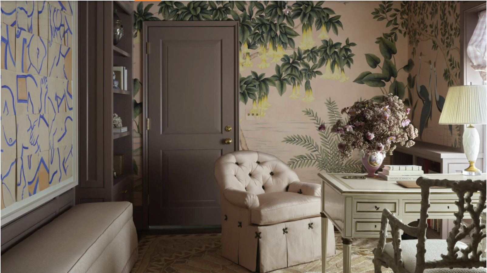 A cozy living room with a beige armchair, a side table with books and a vase of pink and white flowers, a lamp, and a large colorful mural of tropical plants and flowers on the wall.