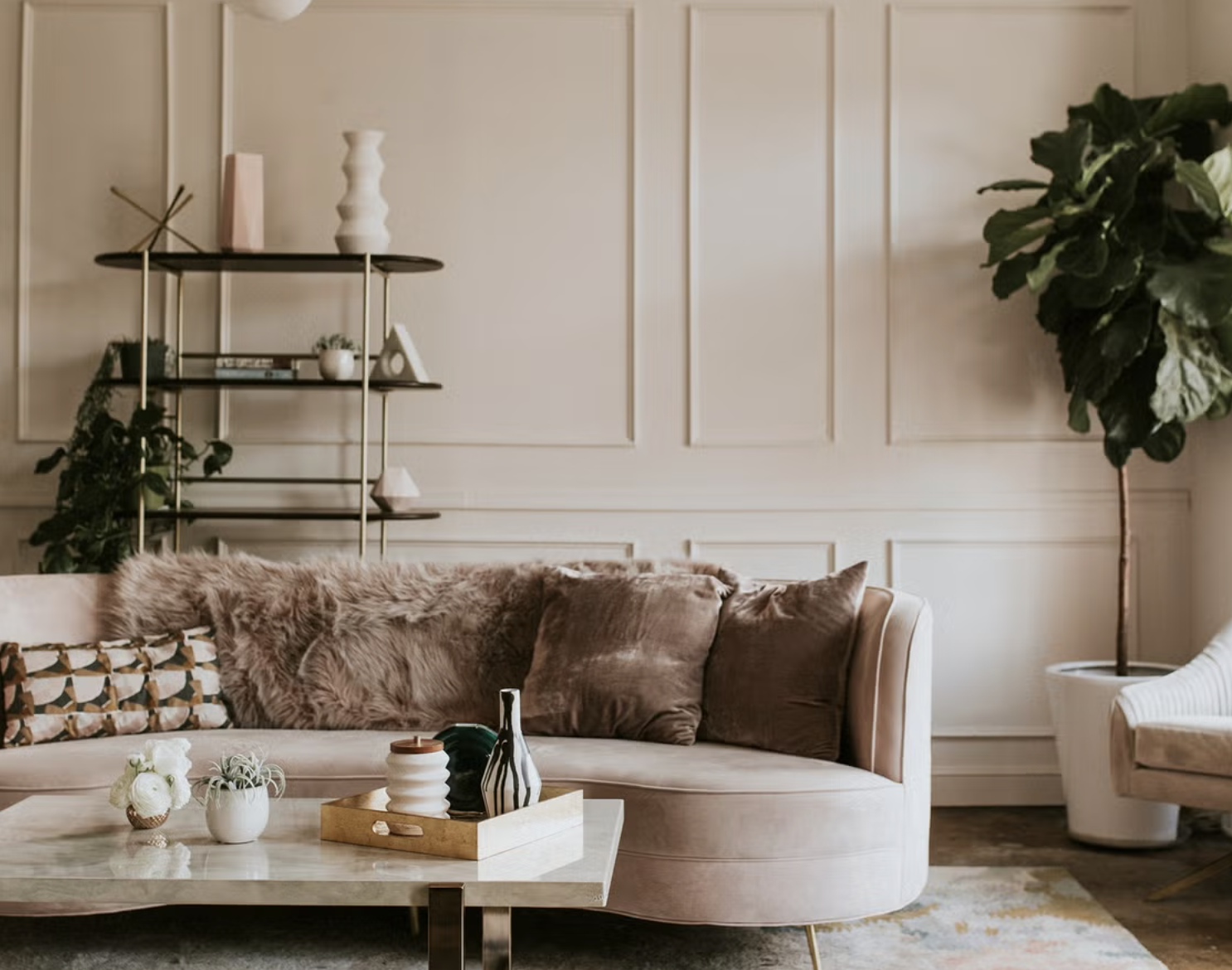 A cozy living room with a beige sectional sofa, decorative pillows, and a fur blanket. A marble coffee table holds small vases and a tray. In the background, there is a black-and-gold shelving unit with decorative items, and a large potted plant on the right side of the room.