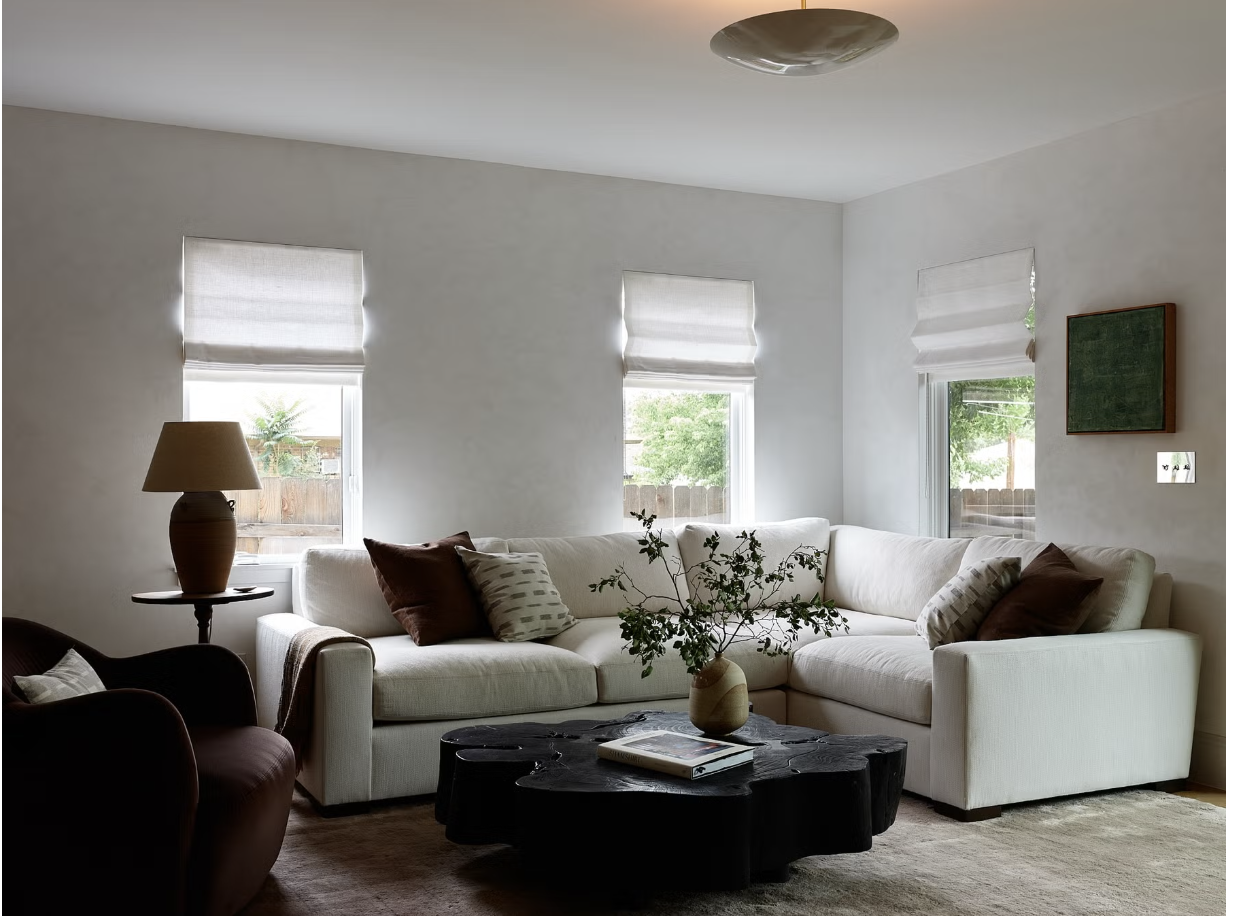 Living room with a white sectional sofa decorated with brown and patterned pillows, a black coffee table with books and a vase with branches, a beige armchair, a table lamp on a side table, three windows with white blinds, and a light-colored wall with a small framed green chalkboard.