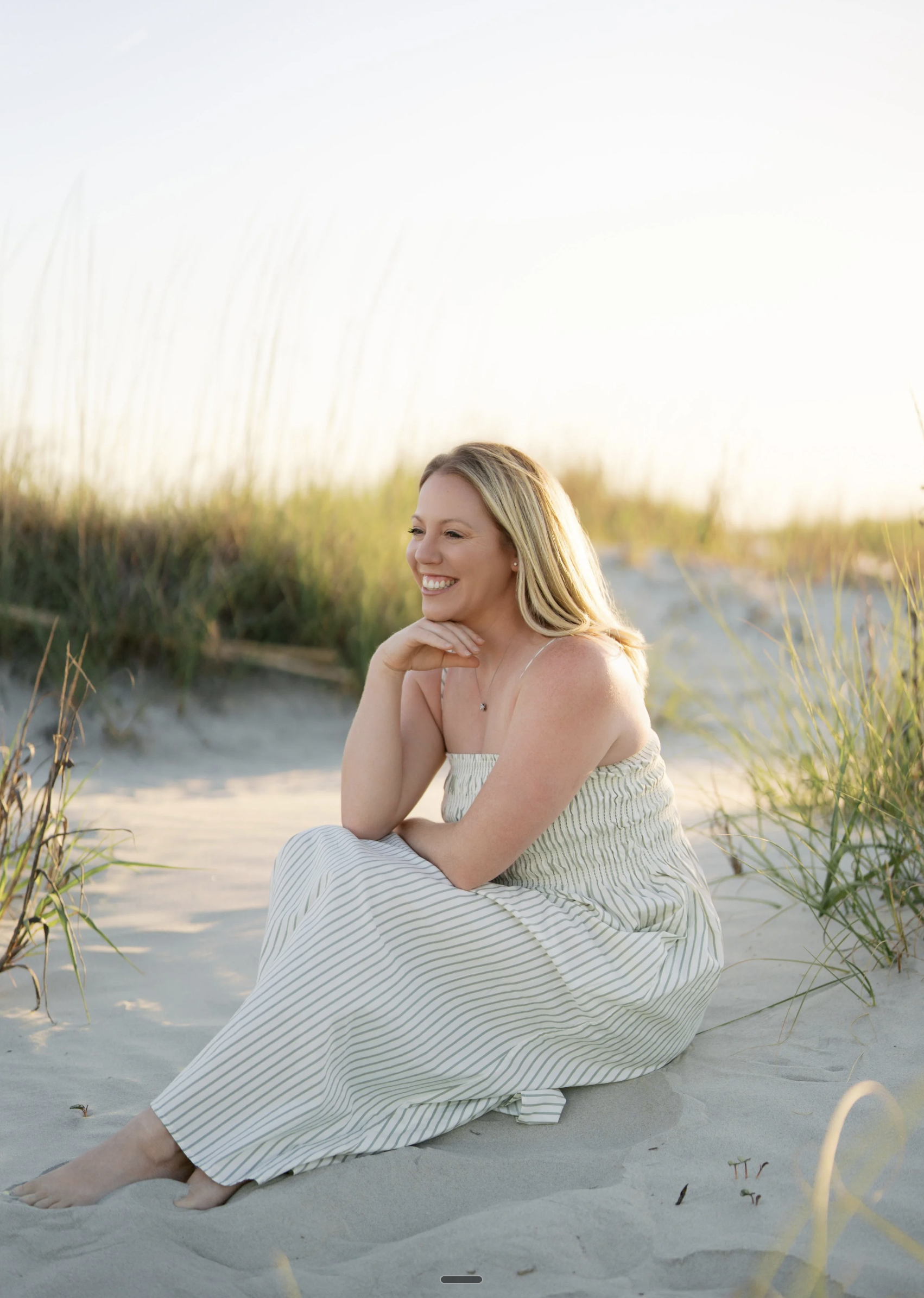 A woman sitting on the sand at the beach, smiling and looking to the side, with tall grass in the background, during sunset.