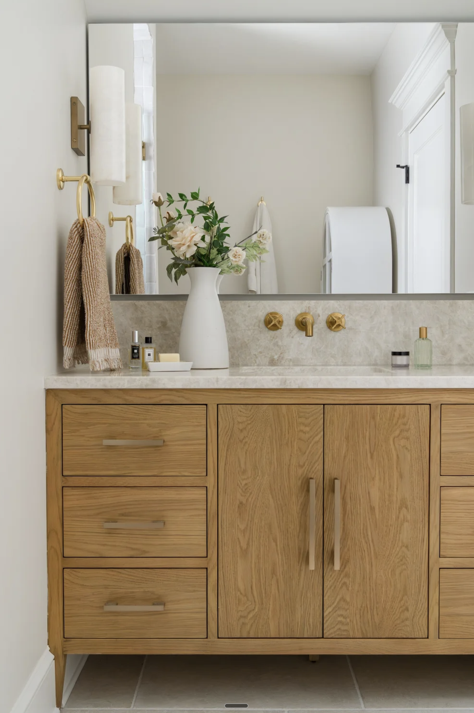 A bathroom vanity with a wooden cabinet, a marble countertop, a large mirror, and various toiletries and decorative items. There are towels hanging on wall hooks and a vase with flowers on the counter.