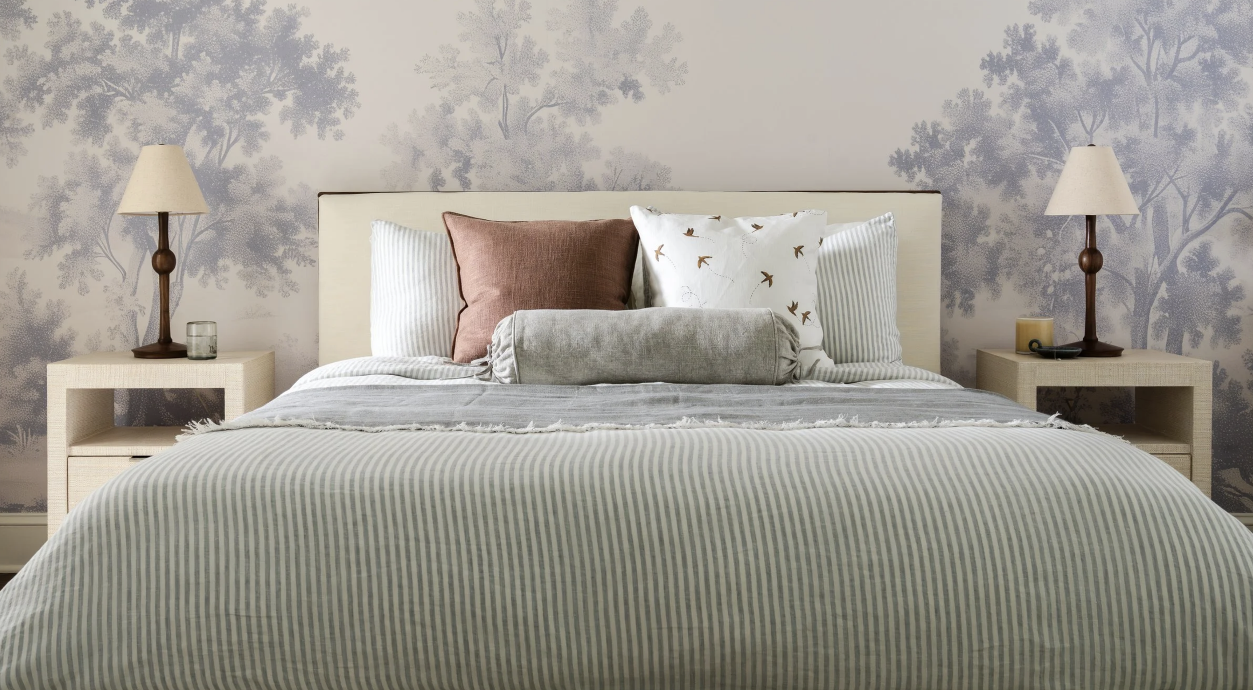 A neatly made bed with striped bedding, decorative pillows including a brown pillow, a pillow with birds, and two striped pillows, flanked by nightstands with lamps and small items, and a wall with a nature-inspired wallpaper pattern.