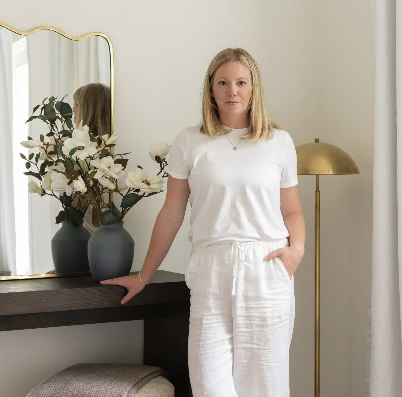 A woman with blonde hair wearing a white t-shirt and white pants standing in a well-lit room with a mirror, vases with white flowers, a gold lamp, and a gray cabinet.