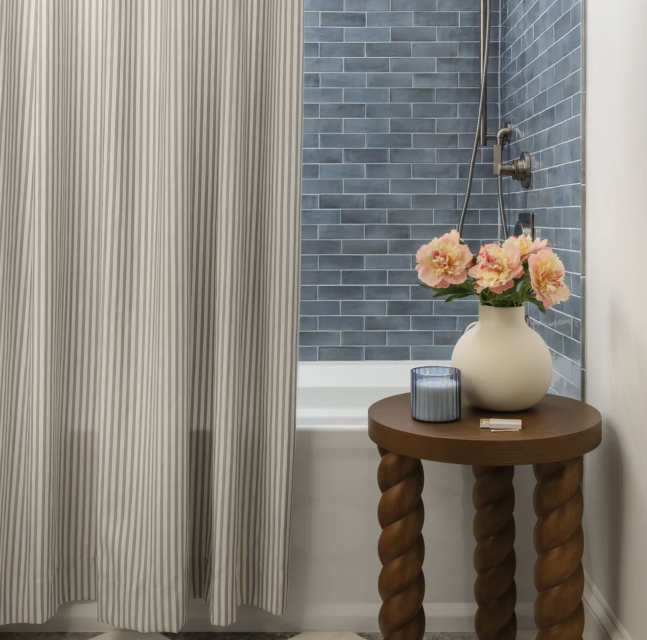 Bathroom with blue subway tile wall, a bathtub, a wooden side table with a white vase of pink flowers, a candle, and a razor.