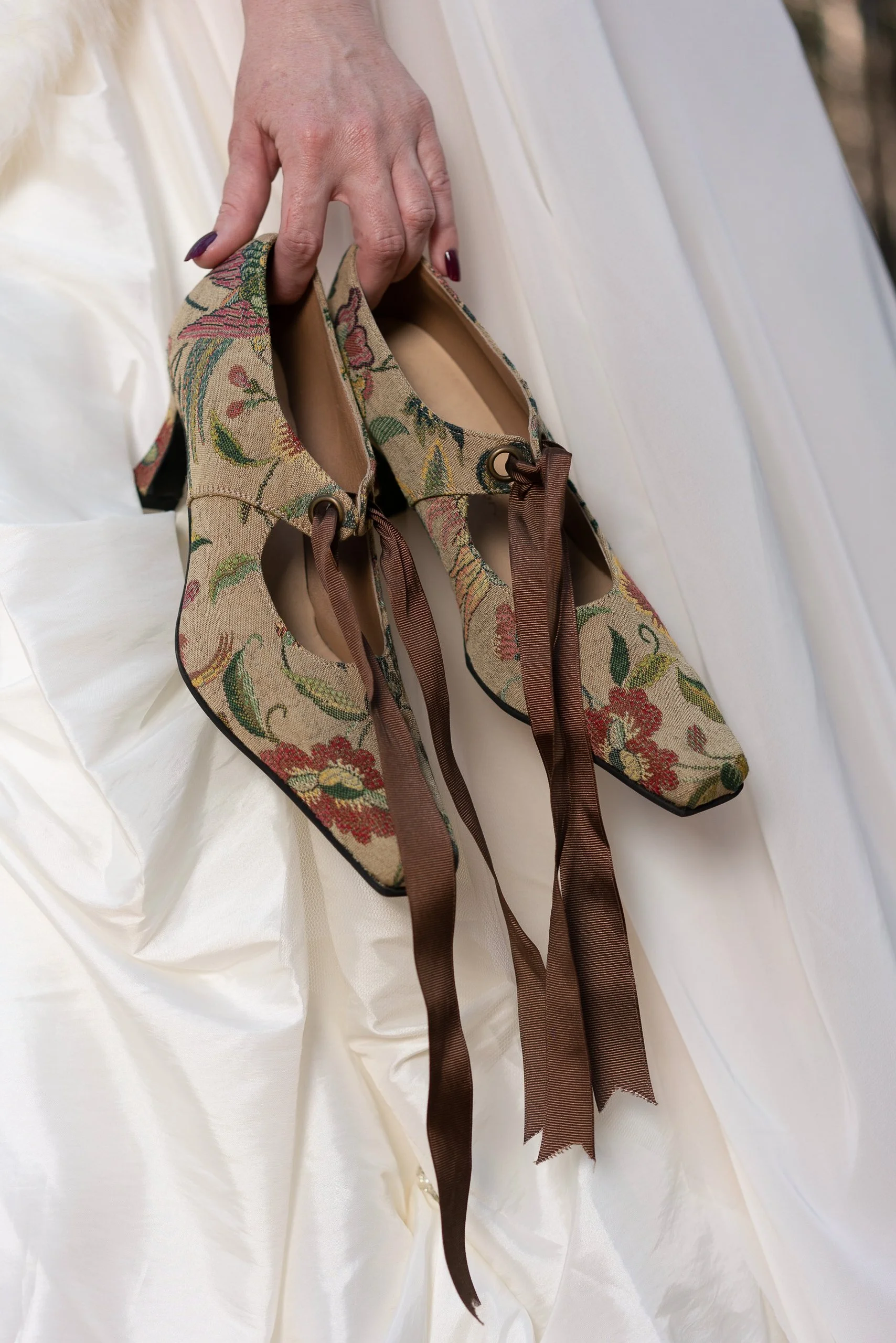Close-up of a person's hand holding a pair of floral-printed ballet flats with brown ribbon laces, set against a white fabric background.