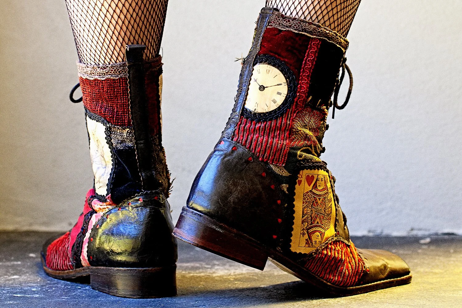 Pair of patchwork cowboy boots with clock and playing card designs, worn over fishnet stockings, on a concrete floor.