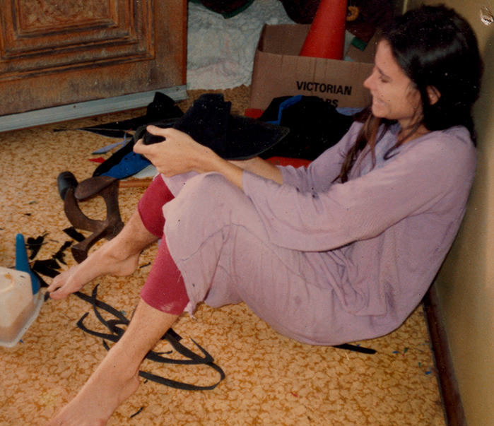 A woman sitting on the floor in a long-sleeve top and pants, holding a pair of black shoes, in a cluttered room with boxes and miscellaneous items.