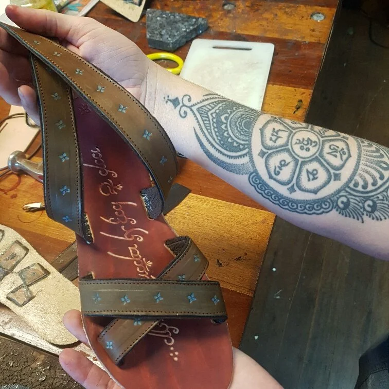 A person's arm with a tattoo holding a brown sandal with blue decorative accents on the straps, on a wooden table with various objects around.