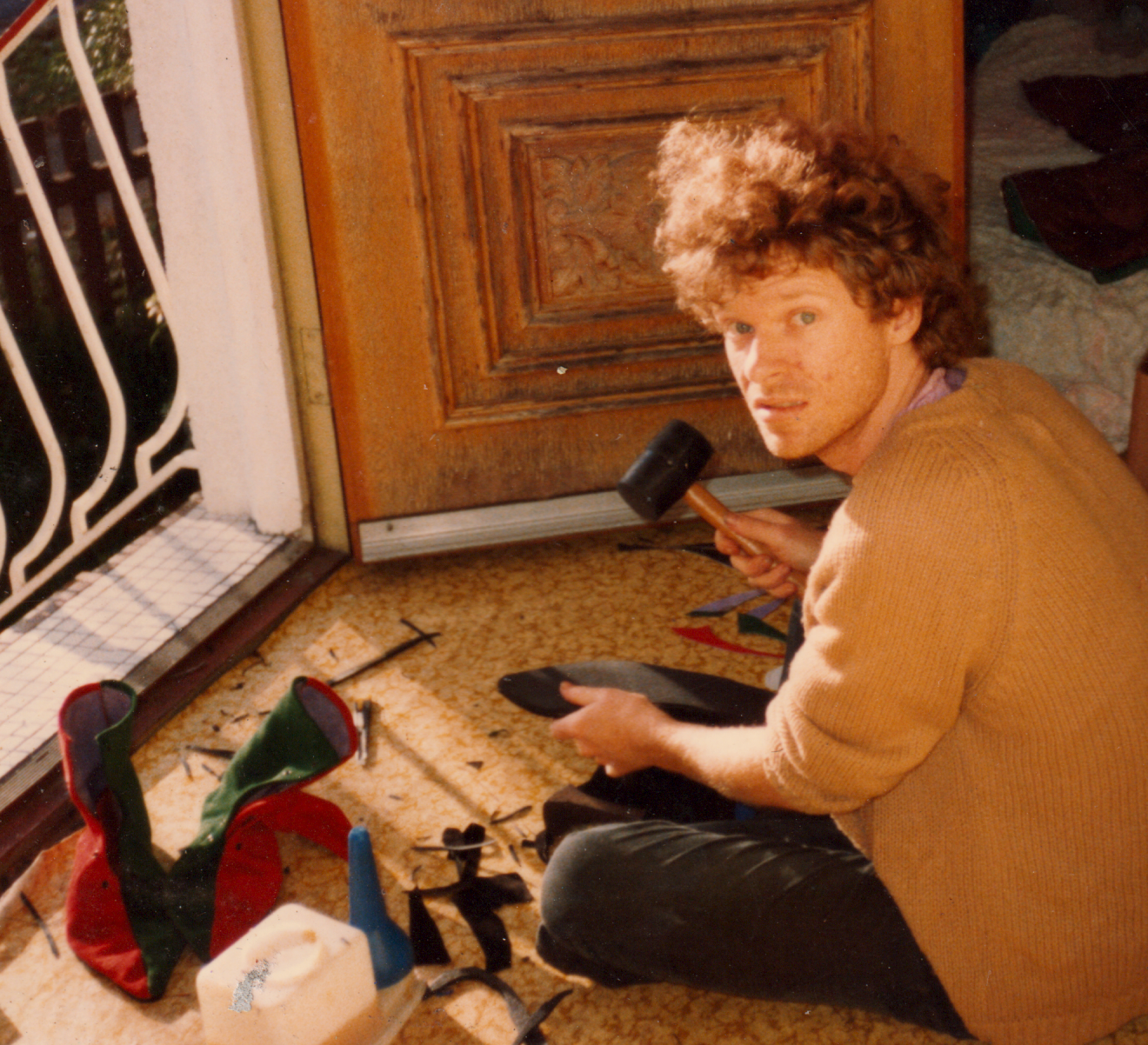 A man with curly hair, wearing a tan sweater, sitting on the floor near a wooden door, holding a hammer and a shoe, surrounded by holiday Christmas stockings, tools, and decorations.