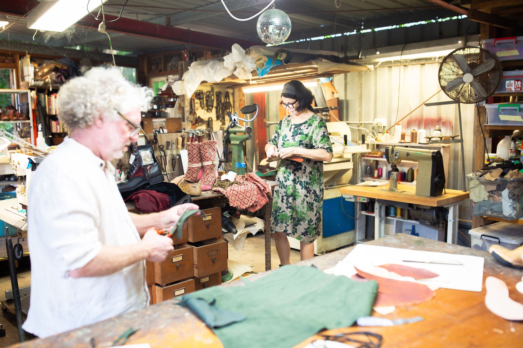 Two women working in a workshop filled with fabric, tools, and equipment. One woman with curly white hair and glasses is focused on a task, while the other woman with dark hair and glasses is looking at a small object. The workshop has shelves, storage bins, and a fan, with natural light coming through the windows.
