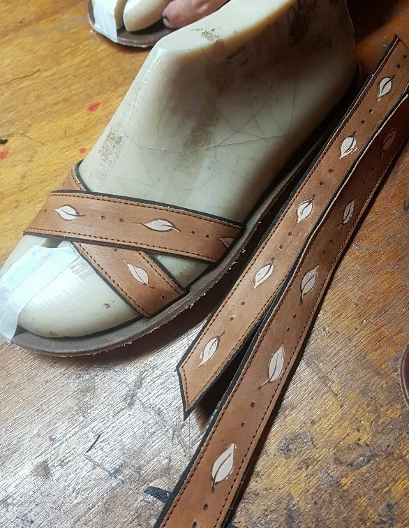A pair of chestnut leather sandals with cut-out leaf patterns on the straps, resting on a wooden surface.