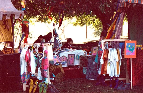 A booth at an outdoor fair or market with various artwork, T-shirts, and crafts for sale under colorful tents and trees.