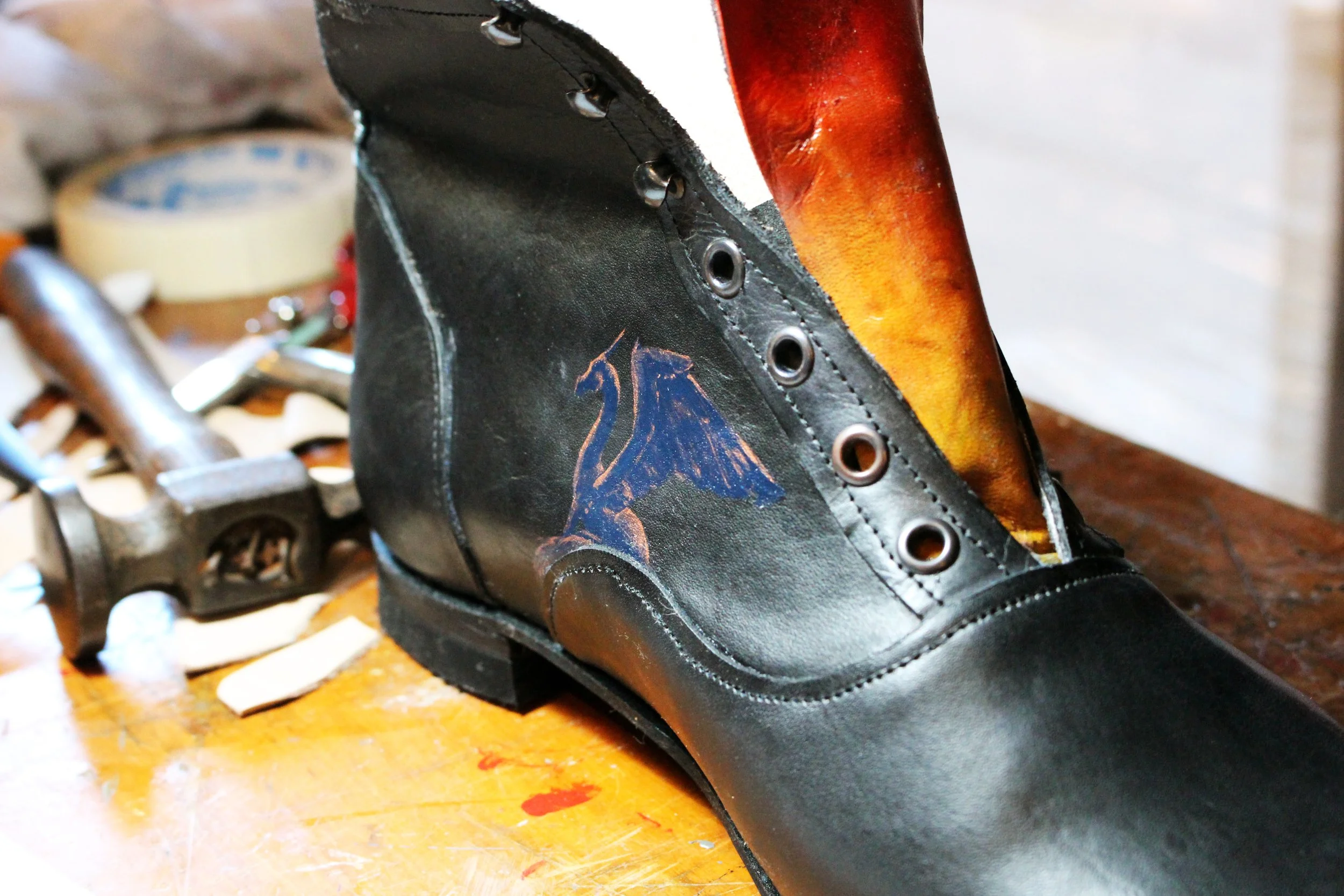 Close-up of a handmade black leather shoe with orange and yellow flame design, showing metal eyelets for laces, on a wooden surface.