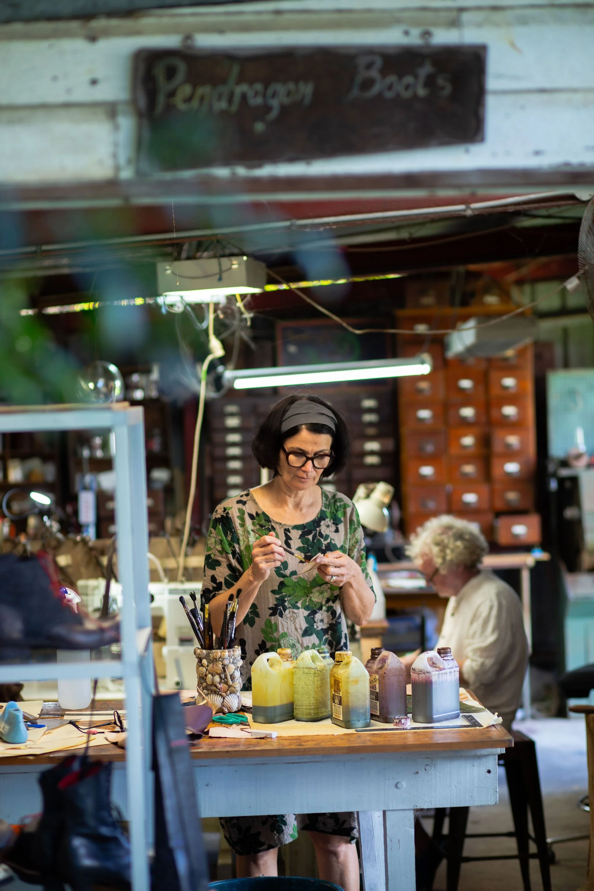 A woman with glasses and dark hair, wearing a floral top, is standing at a work table inside a shop, painting or working on an art project. There are various containers of paint on the table, and an older woman with white hair and glasses is seated in the background, also engaged in an activity.