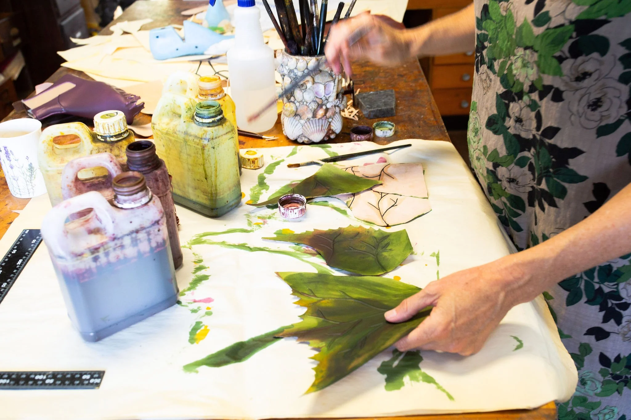Person in a floral dress creating botanical art with green leaves and paint on paper, surrounded by paint bottles and brushes on a wooden table.