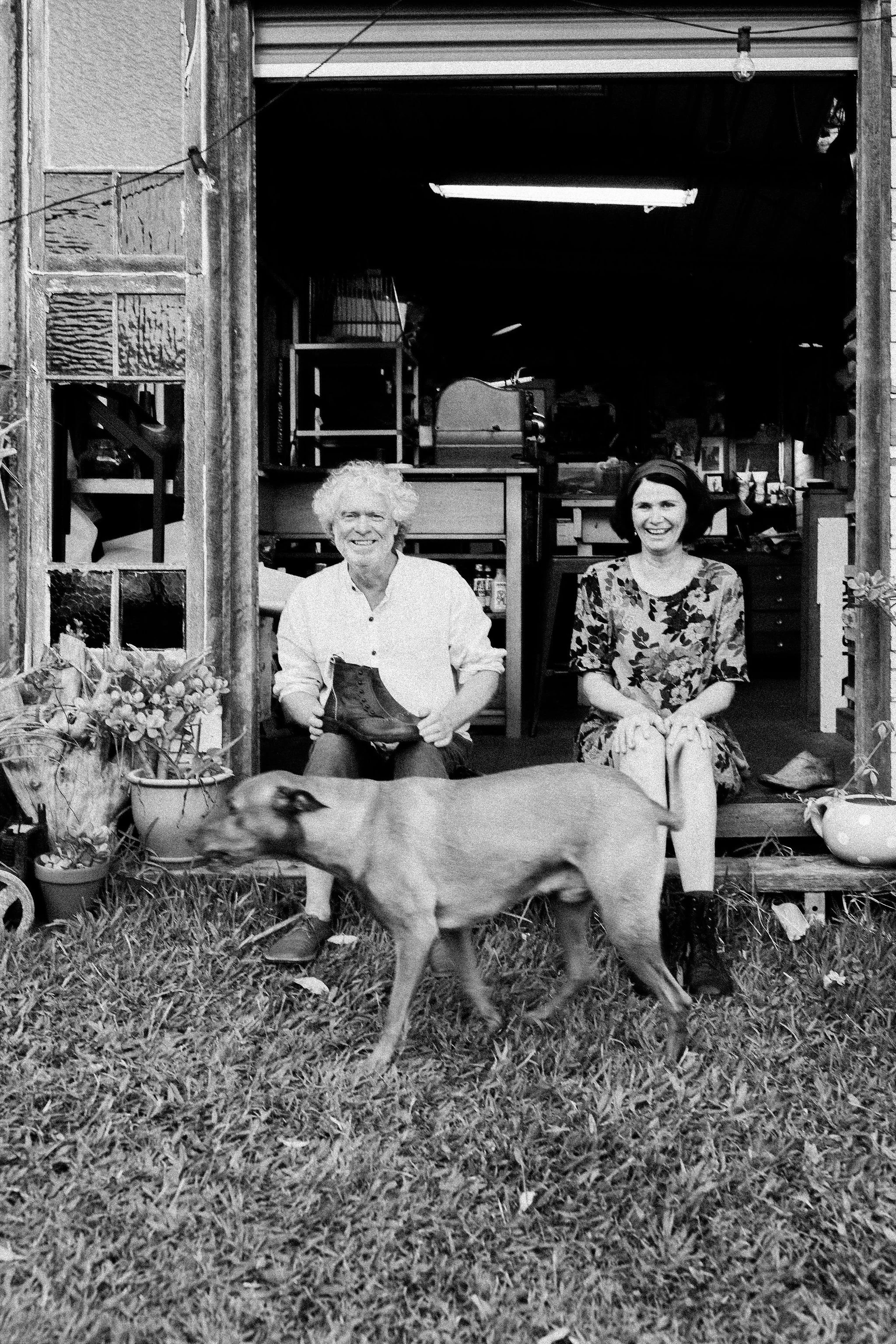 Two women sitting outdoors in front of a building with one of them holding a shoe, with a dog walking in front of them.
