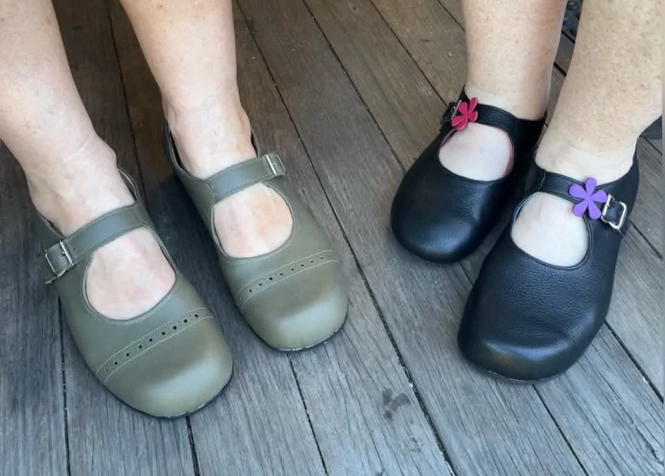 Two people each wearing Mary Jane style shoes they have made in our 3-day shoemaking workshop. One person wears olive green shoes, the other wears black shoes with purple and pink flowers.