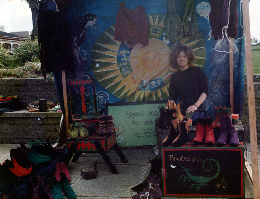 A person with curly hair standing behind a display of handmade shoes and other items at an outdoor market stall. The backdrop features a colorful painted mural with a sun and zodiac symbols.