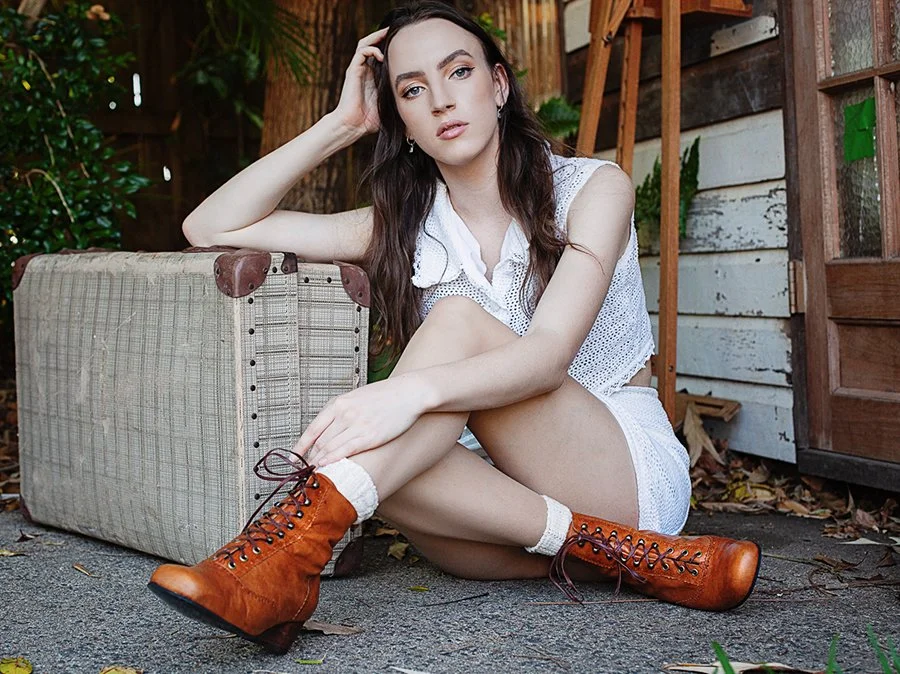 A young woman with long dark hair sits outdoors on the ground, resting her head on her hand. She is wearing a white sleeveless dress, white socks, and brown lace-up boots. There is a vintage suitcase beside her, and a wooden shed and greenery are in the background.