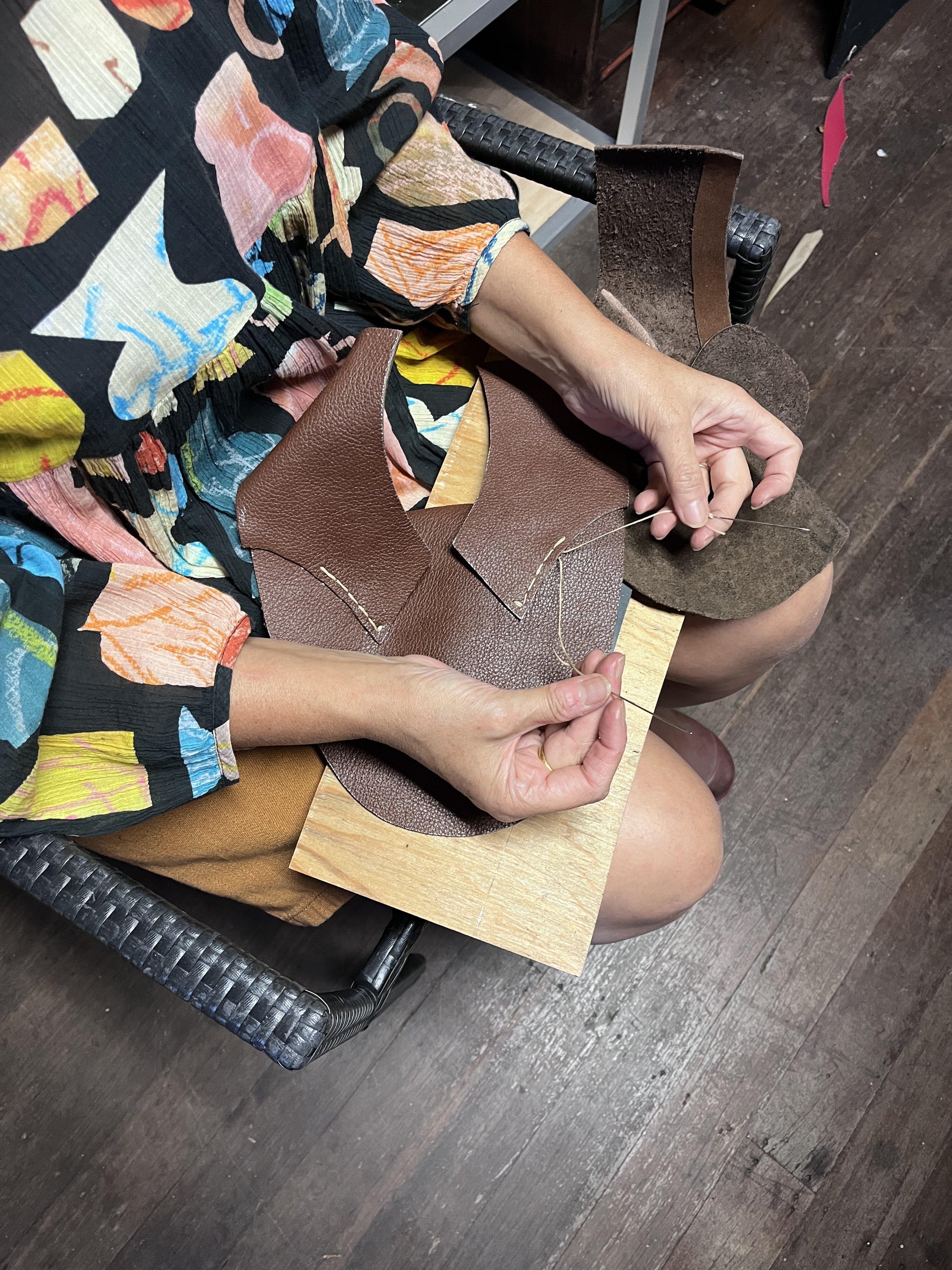 A person sewing brown textured leather on a wooden surface, wearing a colorful abstract patterned dress, seated in a woven chair, dark wood flooring visible in the background.