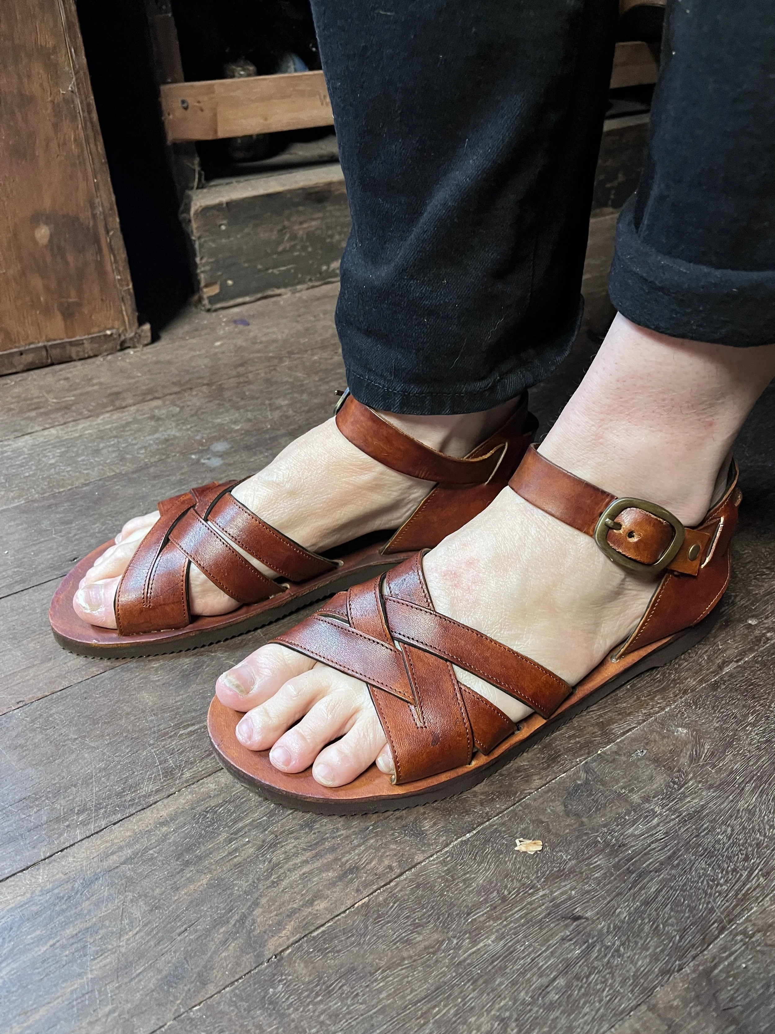 Person wearing brown leather sandals and rolled-up black jeans, standing on a wooden floor.