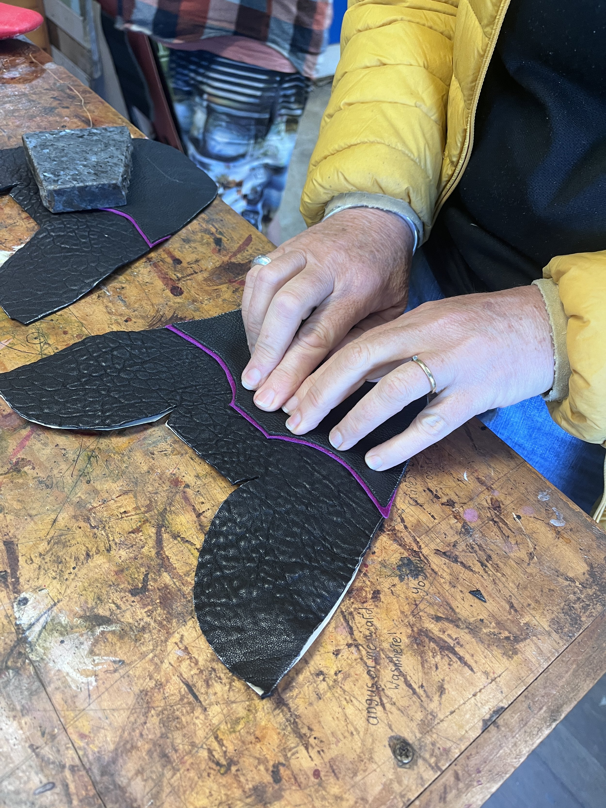 Hands of a person working with black textured leather on a wooden workbench in a workshop, with pieces of leather and a rock-like tool nearby.