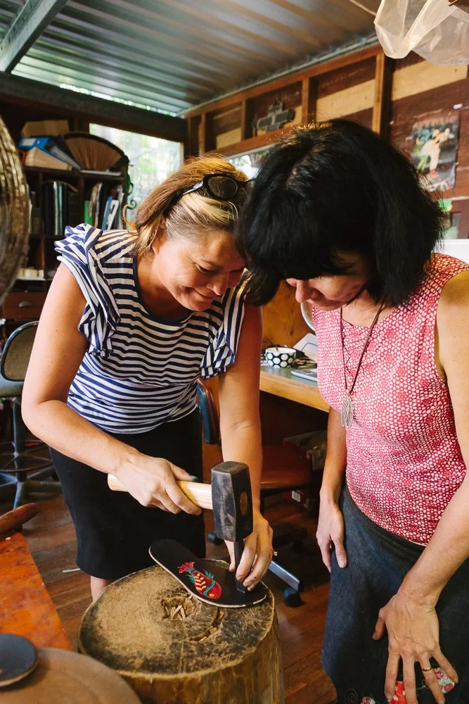 Two women  are engaged in sandal making, with one woman hammering the soles of the sandals.