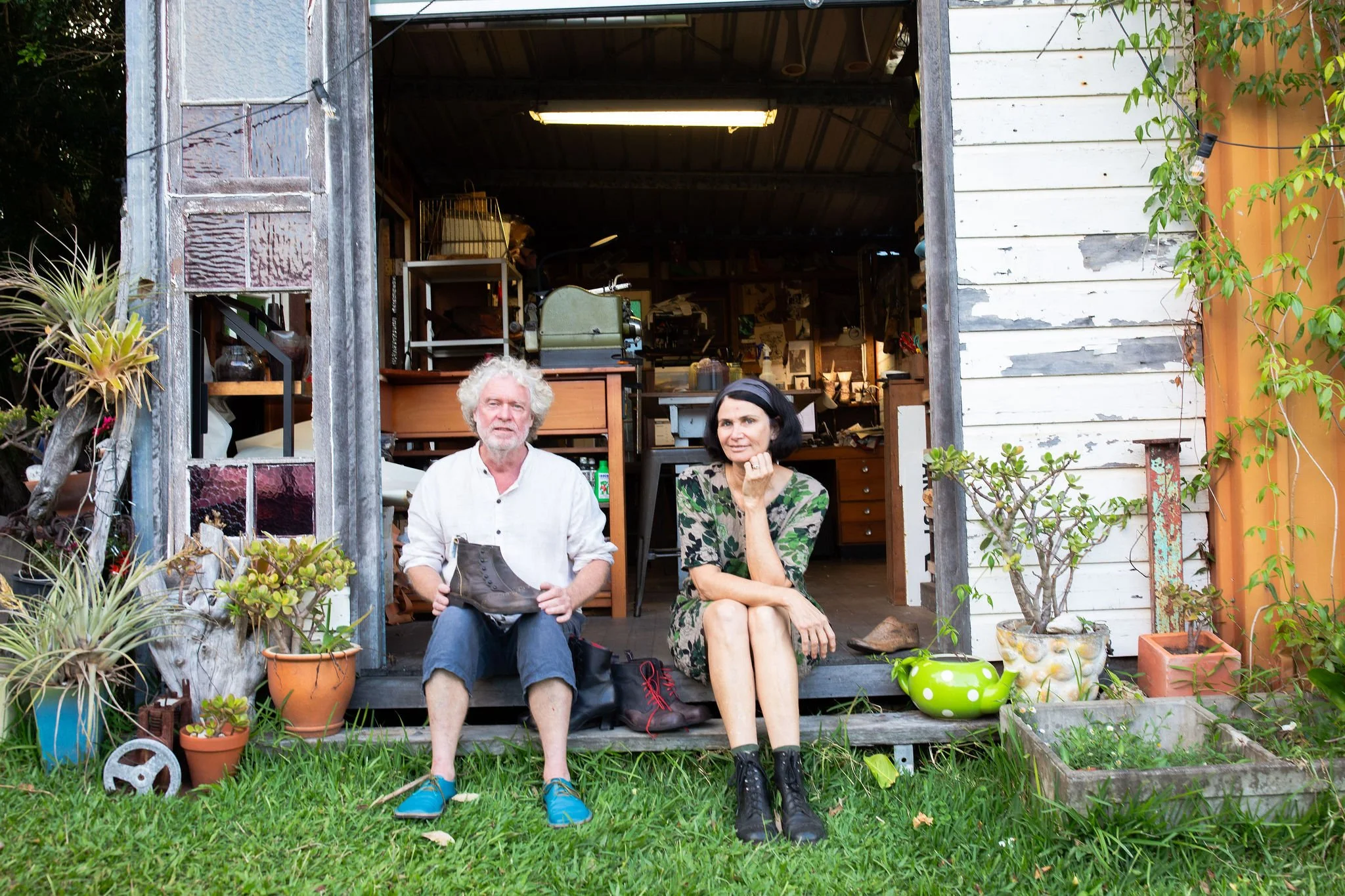 Two people sitting on the porch of a rustic wooden shed, surrounded by potted plants and gardening tools. The man has white curly hair and is holding a shoe, while the woman with black hair is sitting next to him, resting her chin on her hand.