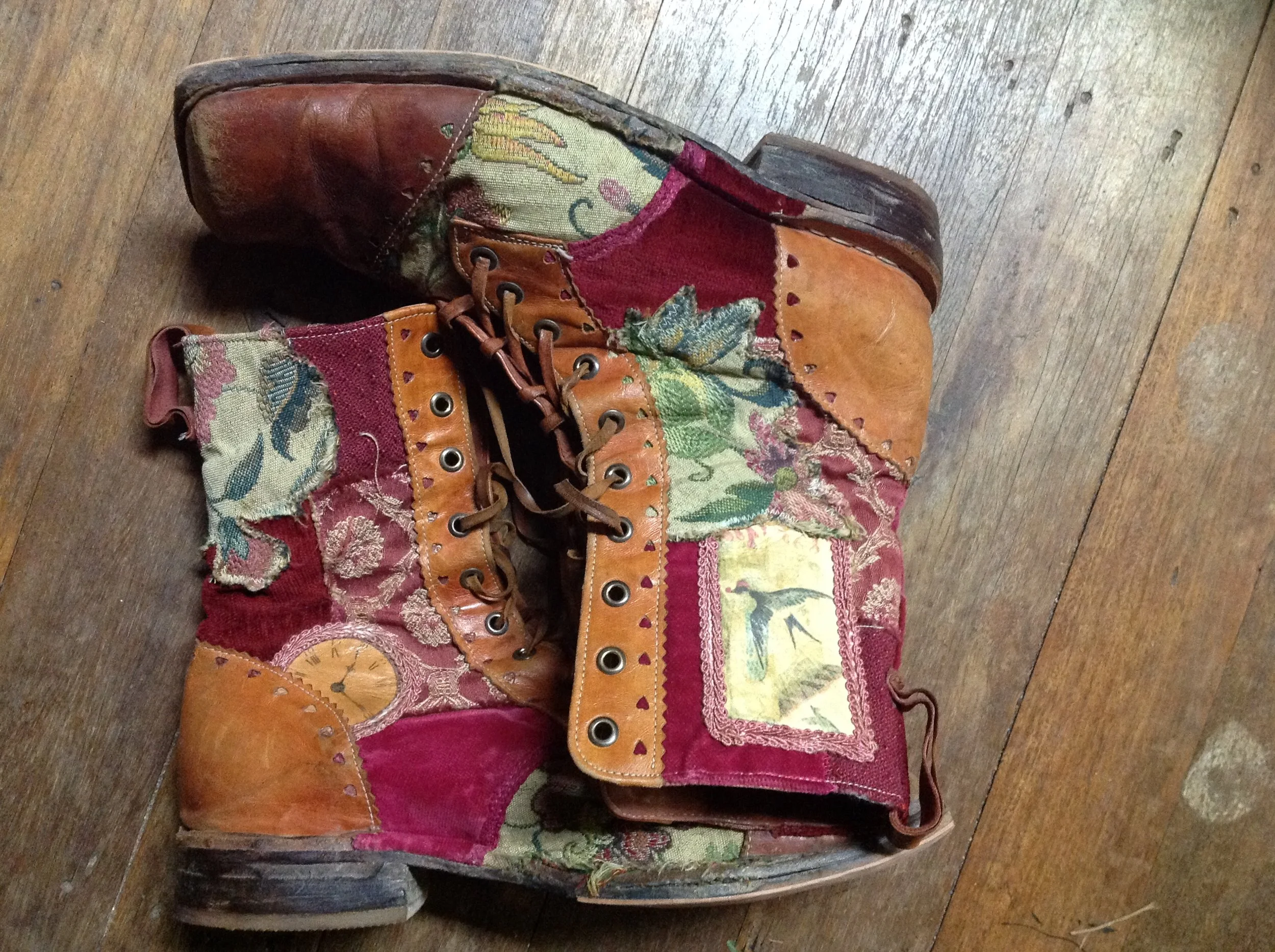 A worn, patched, vintage leather boot with fabric patches and intricate stitching, placed on a wooden floor.