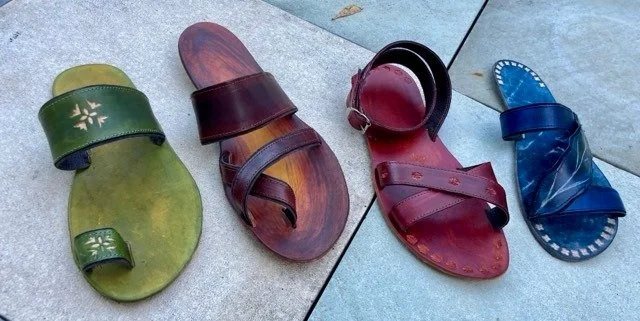 Four pairs of colorful sandals on a tiled surface, in green, brown, red, and blue.