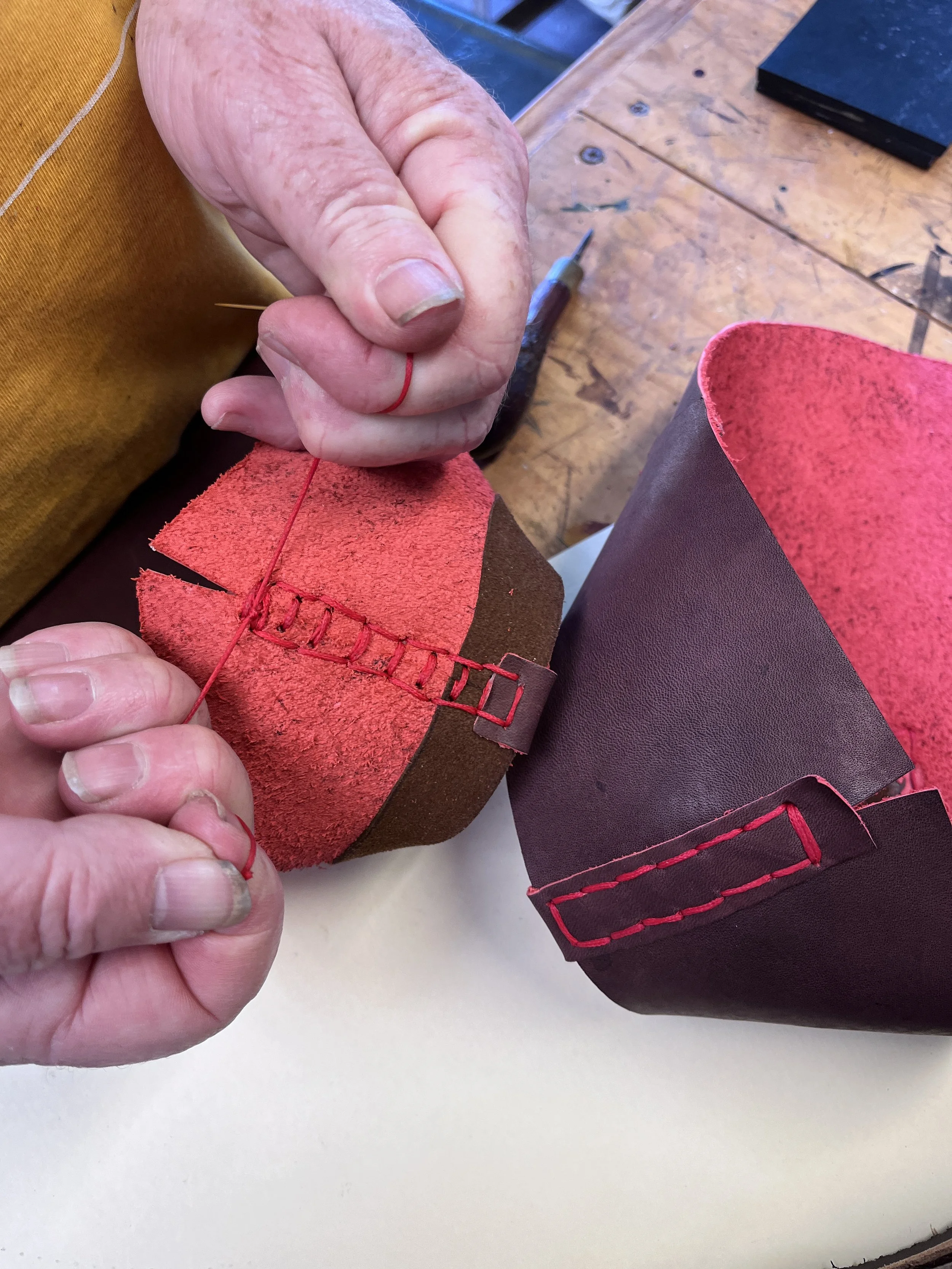 Close-up of a person's hand sewing a piece of red and brown leather with a needle and red thread.