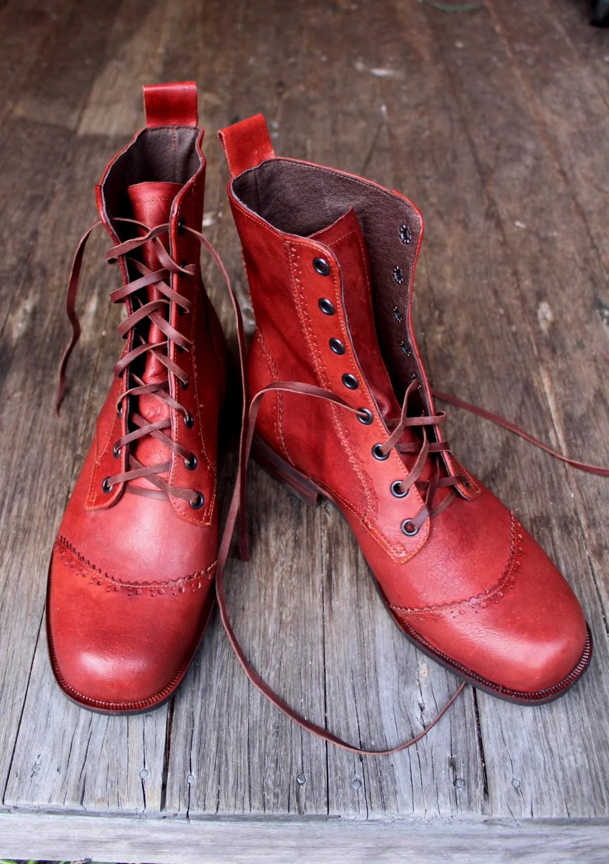 Pair of red leather lace-up ankle boots with black eyelets and a wooden floor background.
