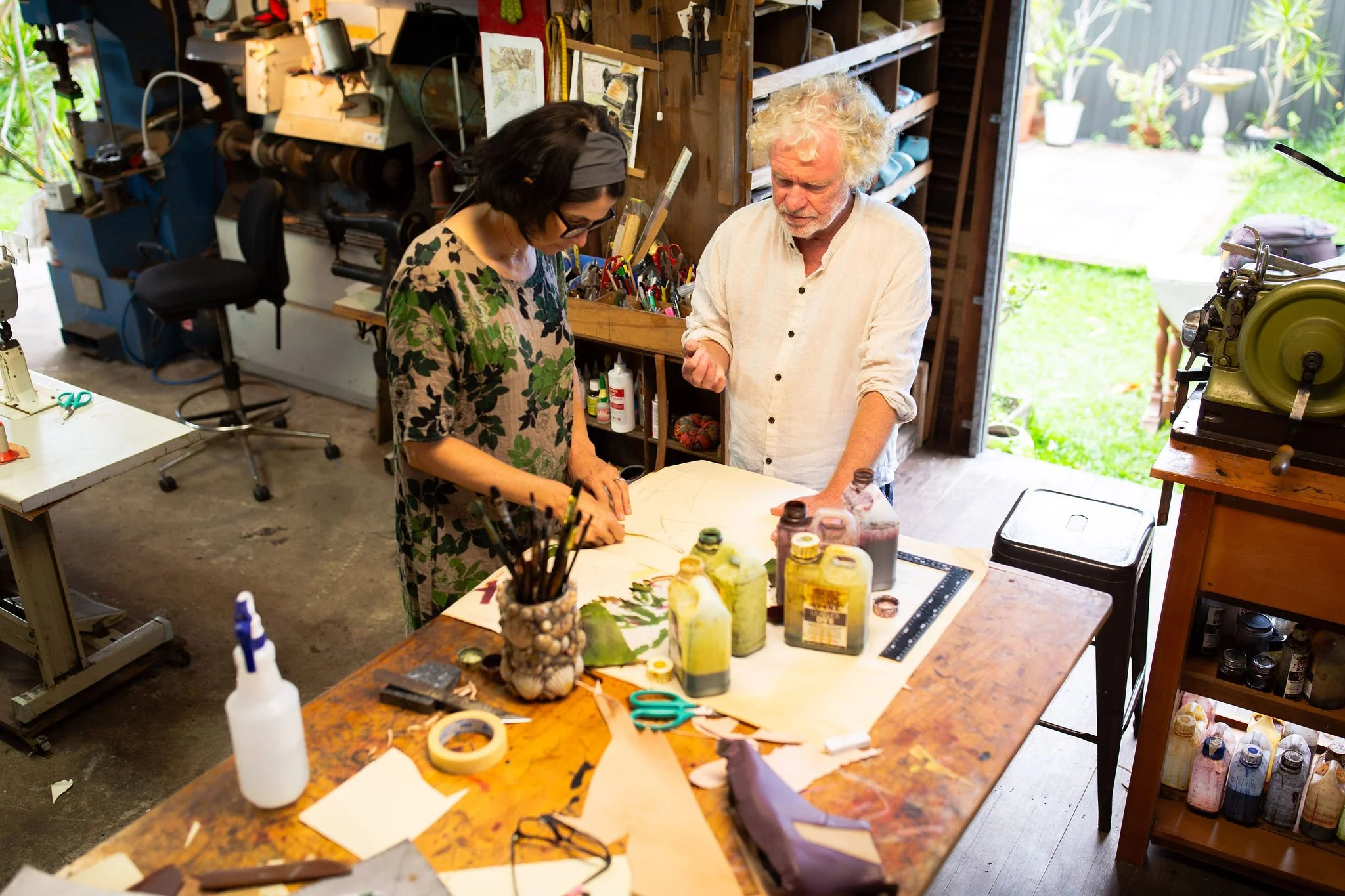 Two people, a woman with dark hair and glasses and an older man with curly white hair, are working together at a cluttered craft or art workspace. They are both observing a large sheet of paper or fabric on a table, which has various paints, brushes, and art supplies around it. The room has shelves with tools and supplies, and large open doors showing greenery outside.