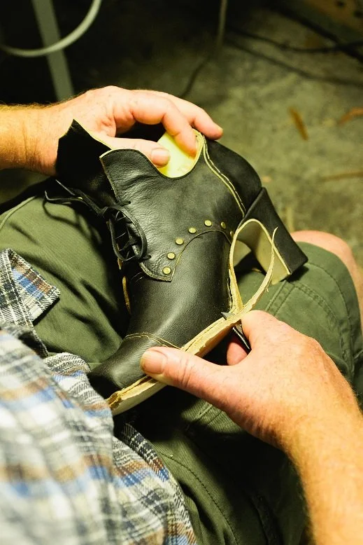 A person sitting down with a plaid shirt, holding a black leather dress shoe in their hands, repairing or polishing it.