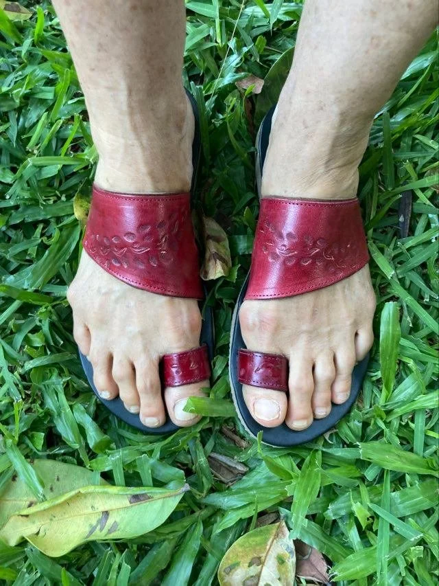 A pair of feet wearing red leather sandals with decorative patterns, standing on green grass with some brown leaves.