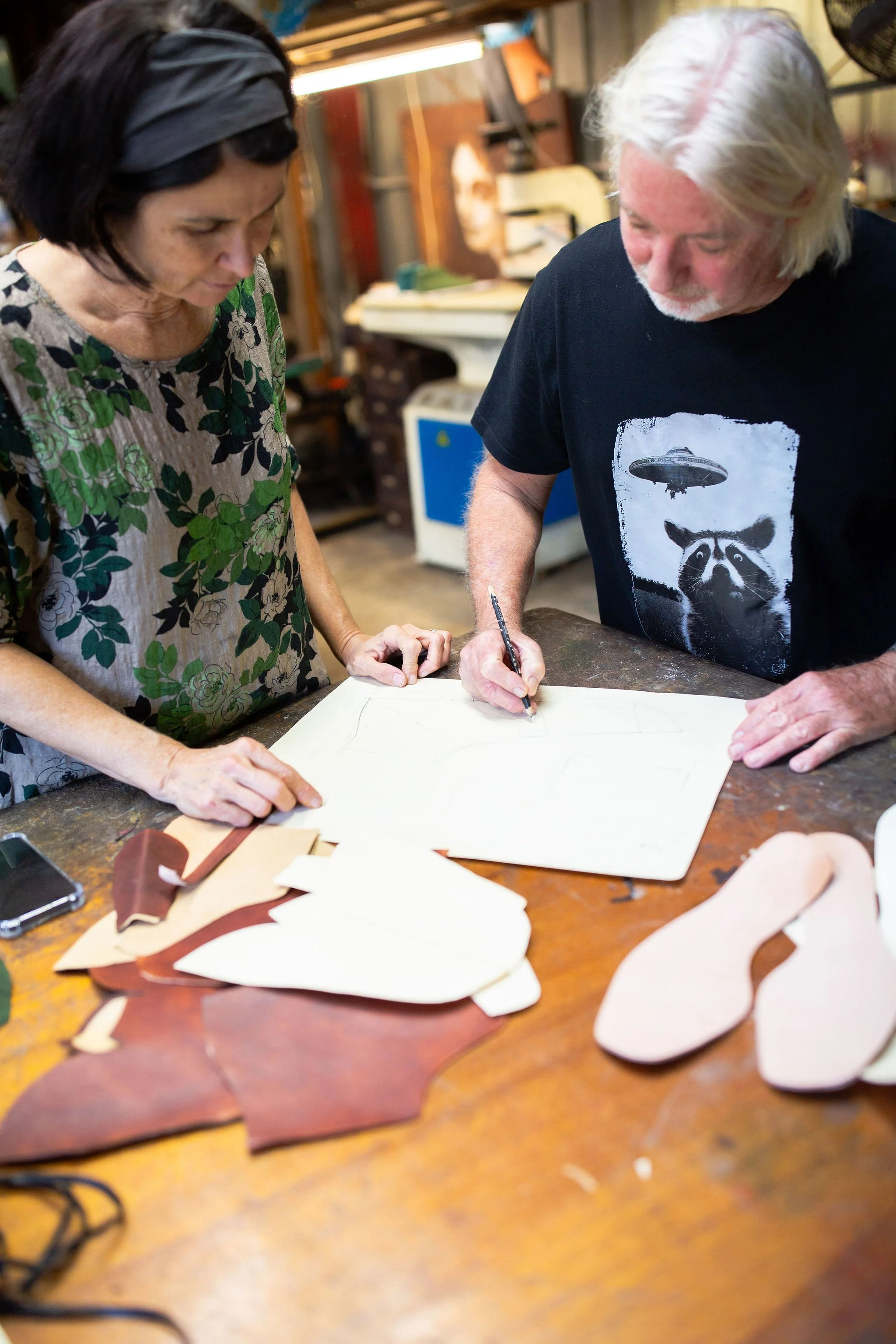 Two people working in a boot making workshop at a wooden table with leather cutouts, in a workshop filled with various tools and materials.